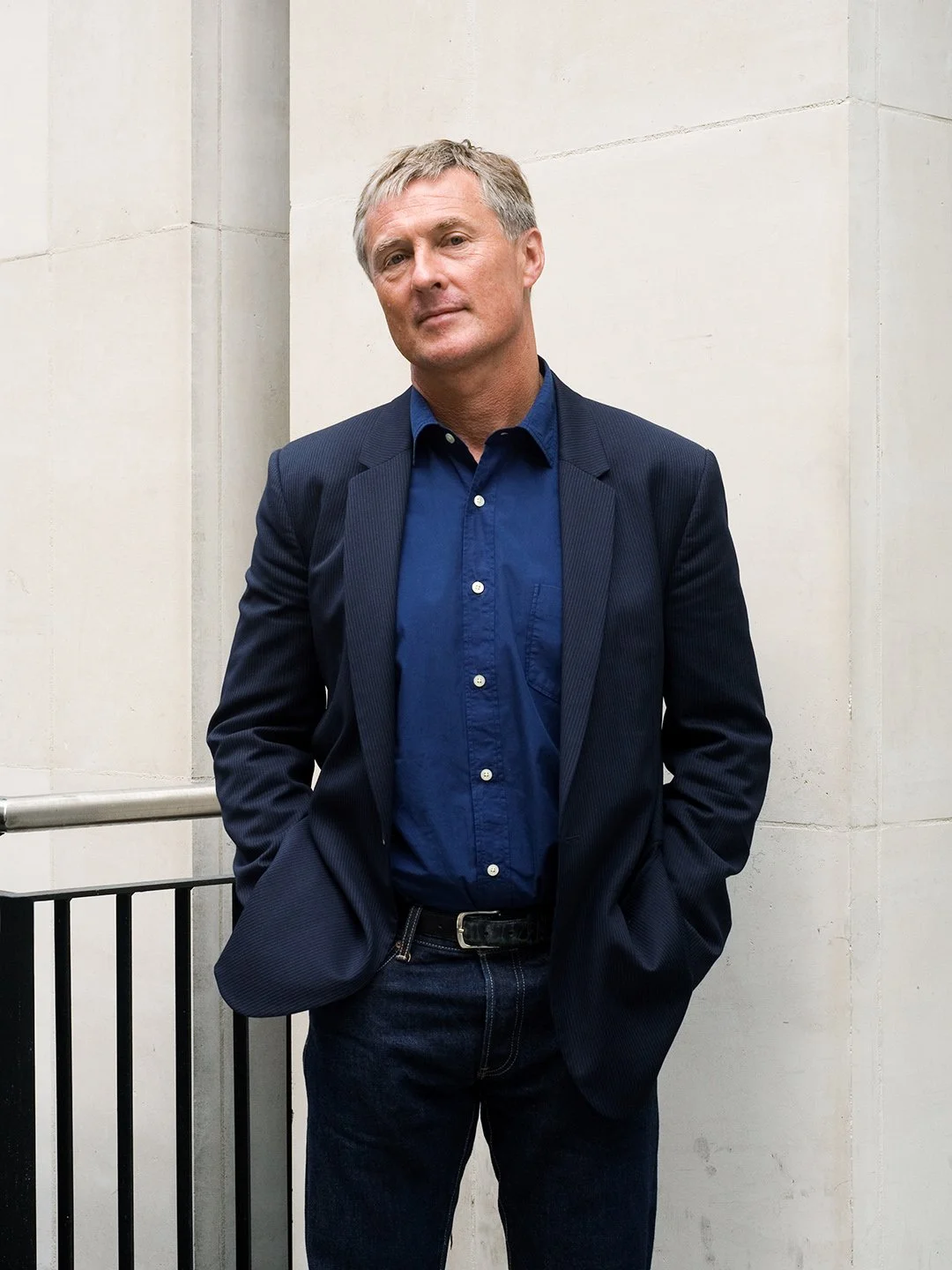 A man with short blonde hair wearing a dark blue blazer, blue button-up shirt, and dark jeans standing against a light-colored wall with his hands in his pockets.