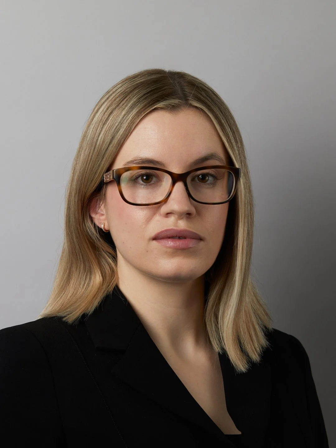A professional woman with shoulder-length blonde hair, wearing glasses and a black blazer, posing against a plain gray background.