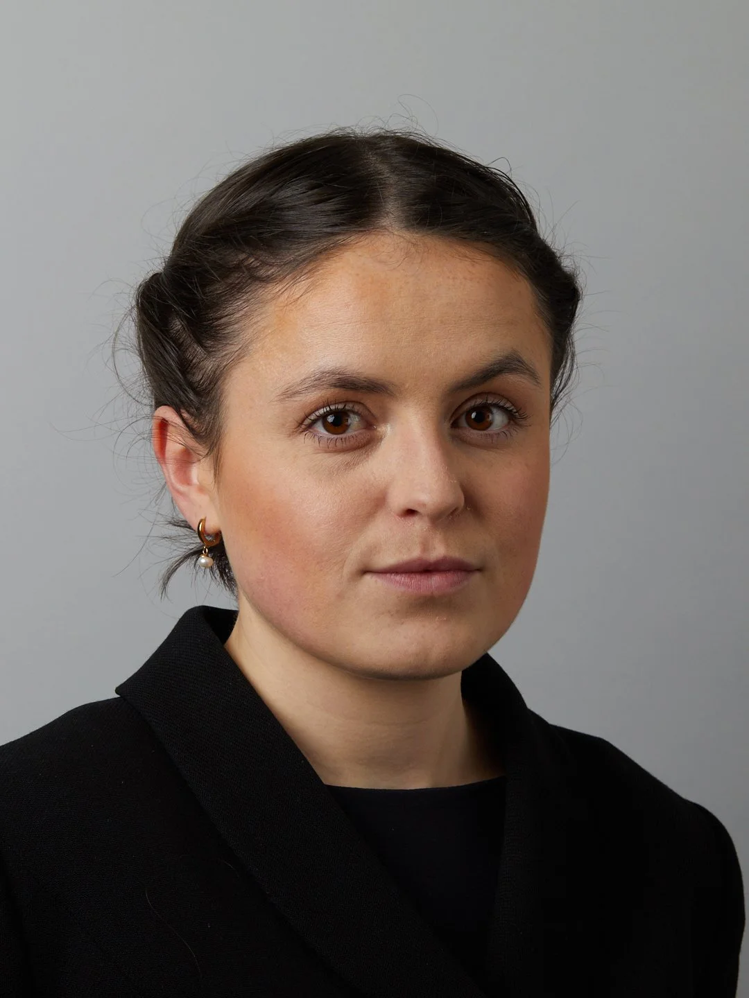 A woman with short dark hair, wearing a black jacket and small hoop earrings with pearls, posing against a plain grey background.