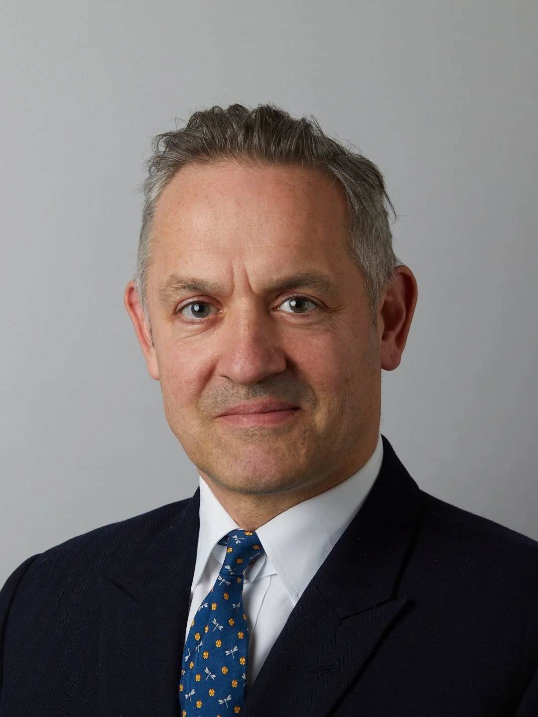 A professional headshot of a middle-aged man with short gray hair, blue eyes, wearing a dark suit, white shirt, and a blue tie with yellow and white patterns, against a plain light gray background.