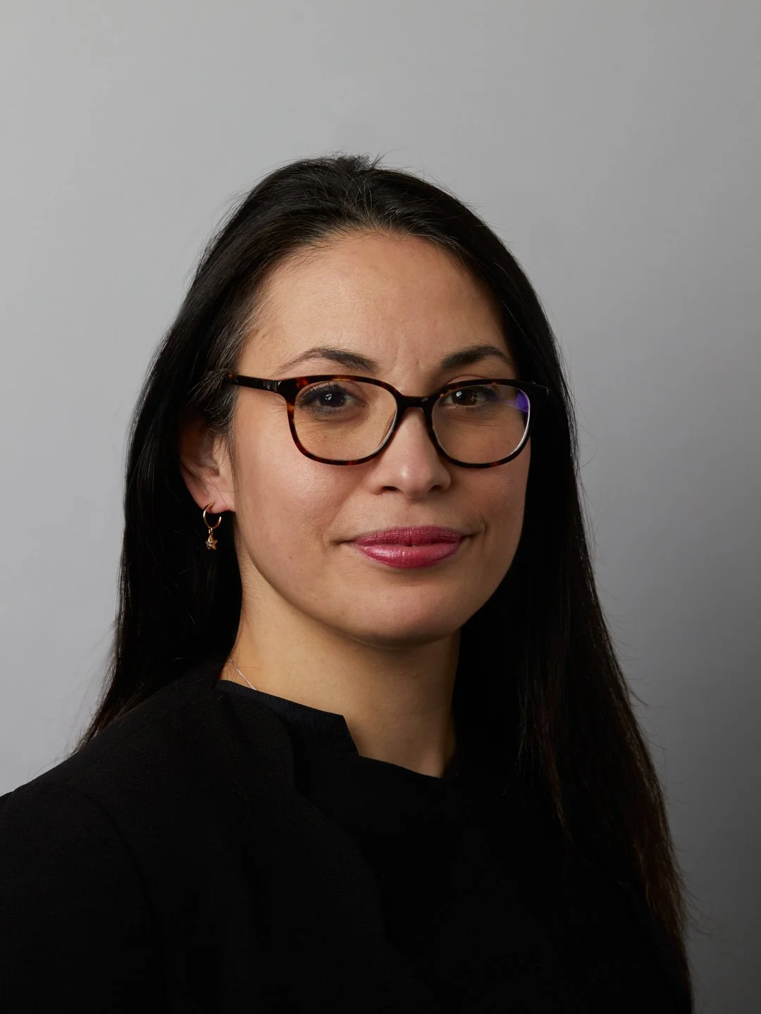 Professional portrait of a woman with long dark hair, wearing glasses and earrings, against a neutral background.