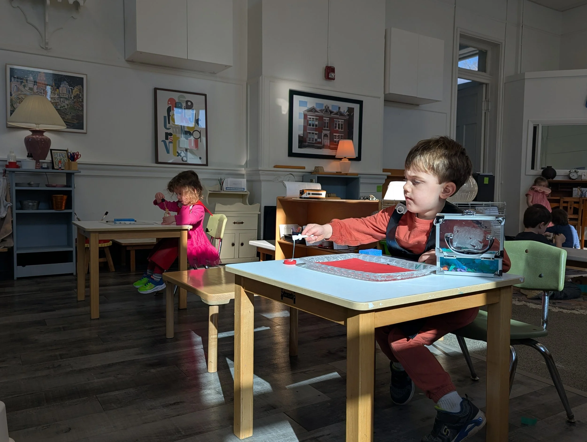 Children in a classroom engaged in activities at their tables, with sunlight streaming in through the window.
