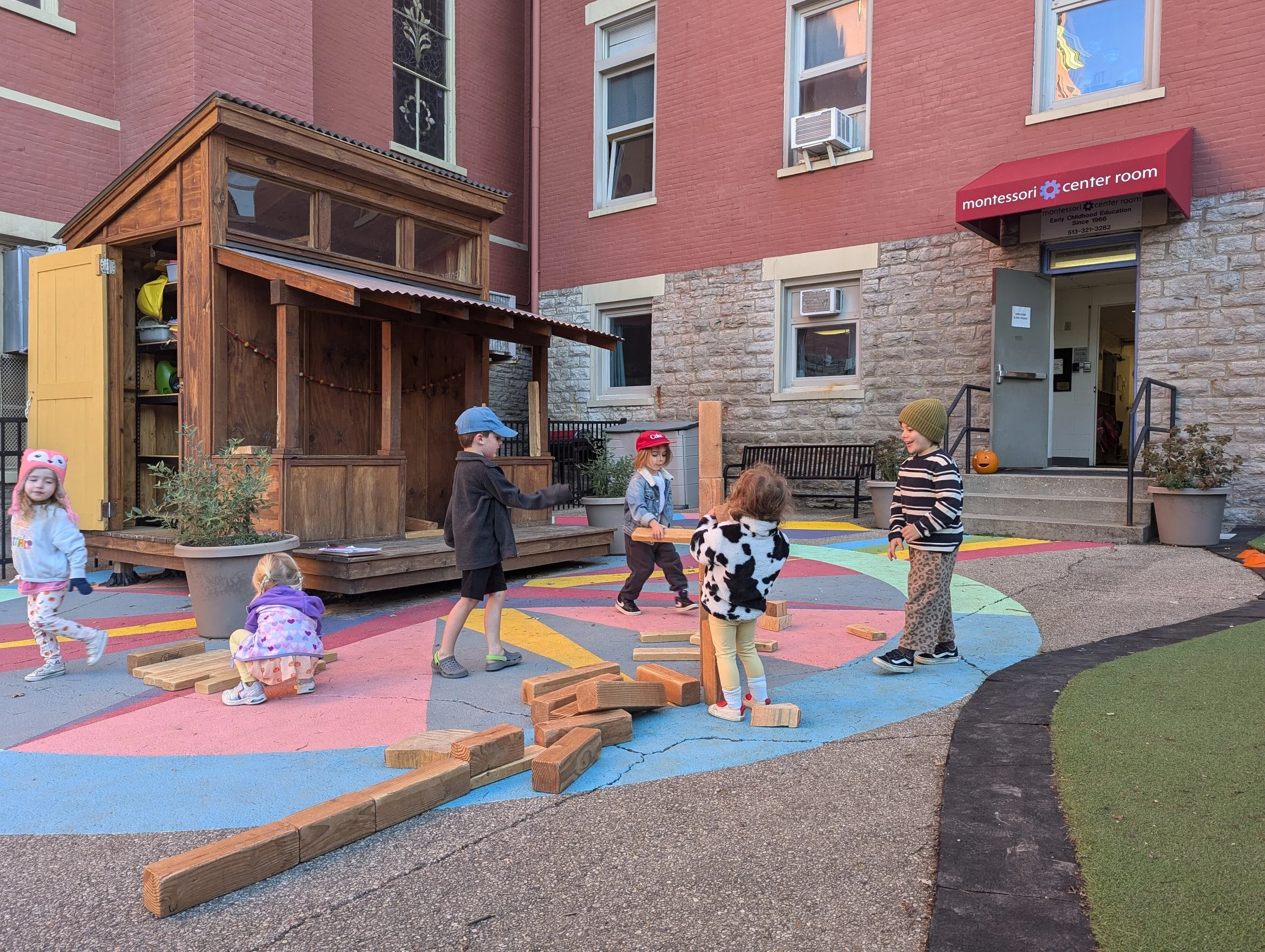 Children playing outside a preschool or daycare center, with a small wooden stage, colorful playground markings on the pavement, and an entrance to the building labeled 'Montessori Center Room' with an autumn pumpkin decoration.
