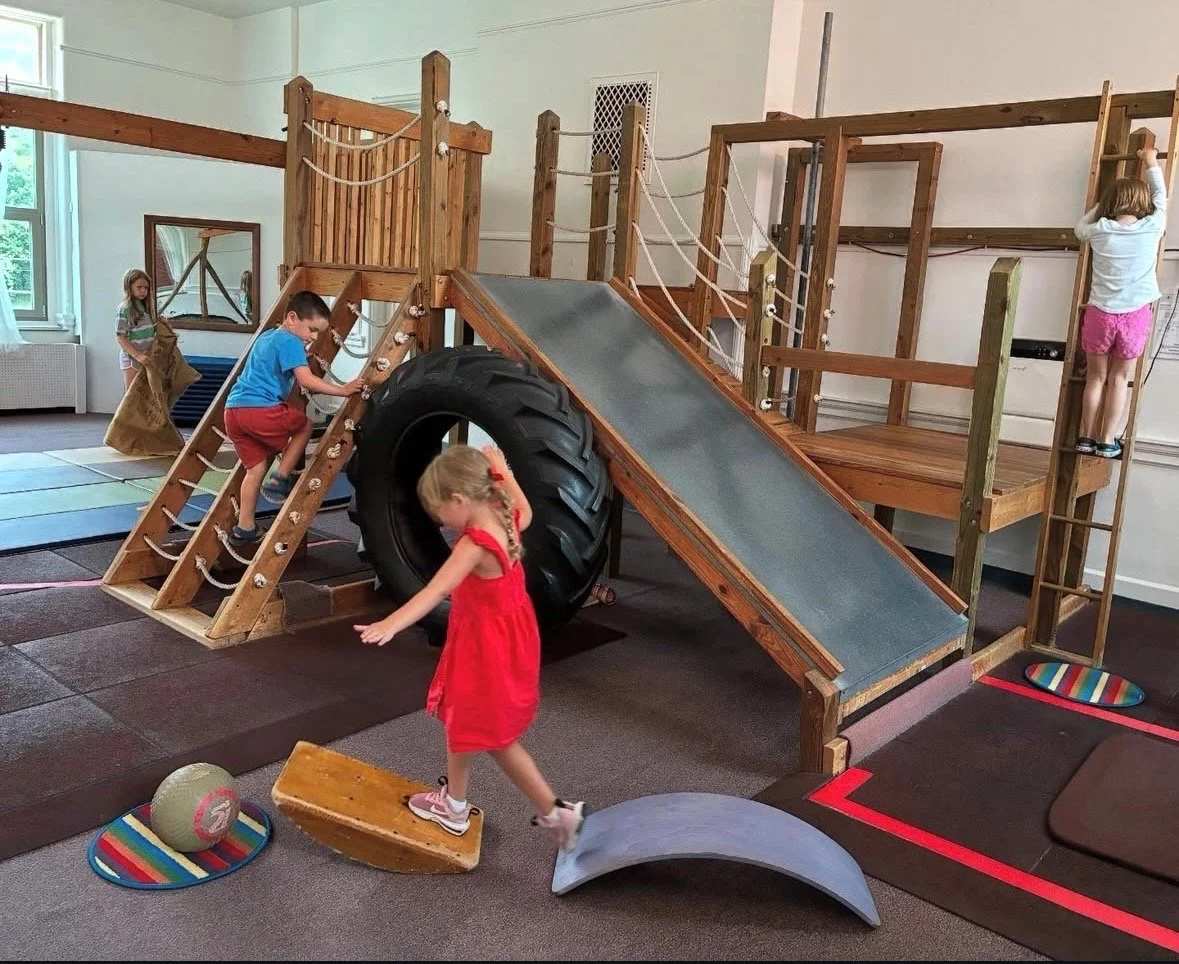 Children playing on an indoor wooden play structure with a slide, tire tunnel, and climbing areas.