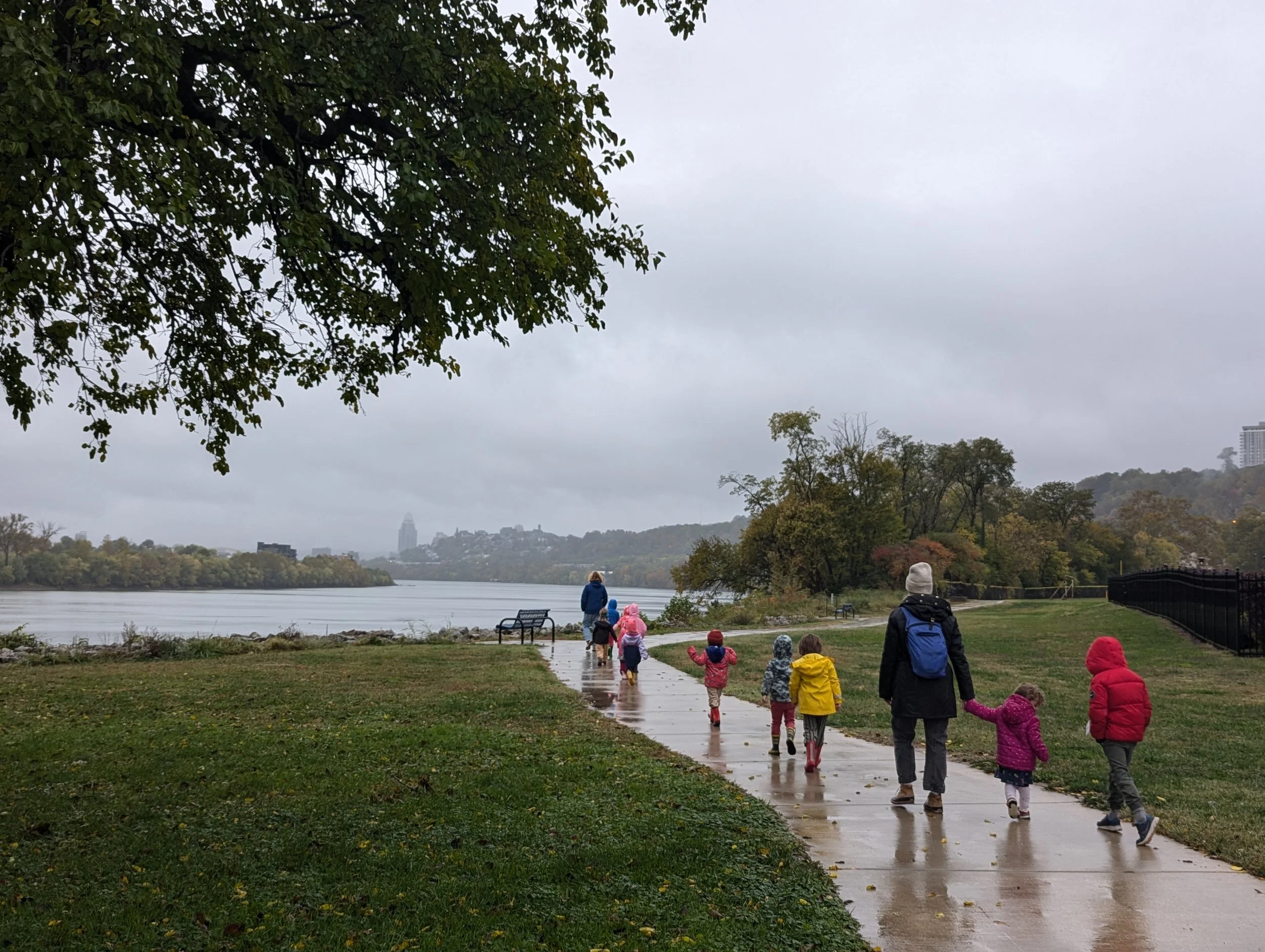 A group of children and two adults walking on a wet sidewalk along a river on a cloudy day. The children are dressed in colorful jackets and hats. The scene is surrounded by trees and greenery, with city buildings visible in the distance.