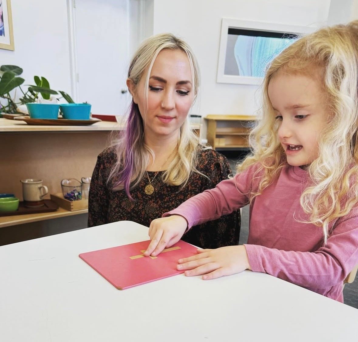 A woman with blonde hair and a girl with curly blonde hair are sitting at a white table. The girl is pointing at a red book, and the woman is observing. There are shelves with mugs, bowls, and a plant in the background.