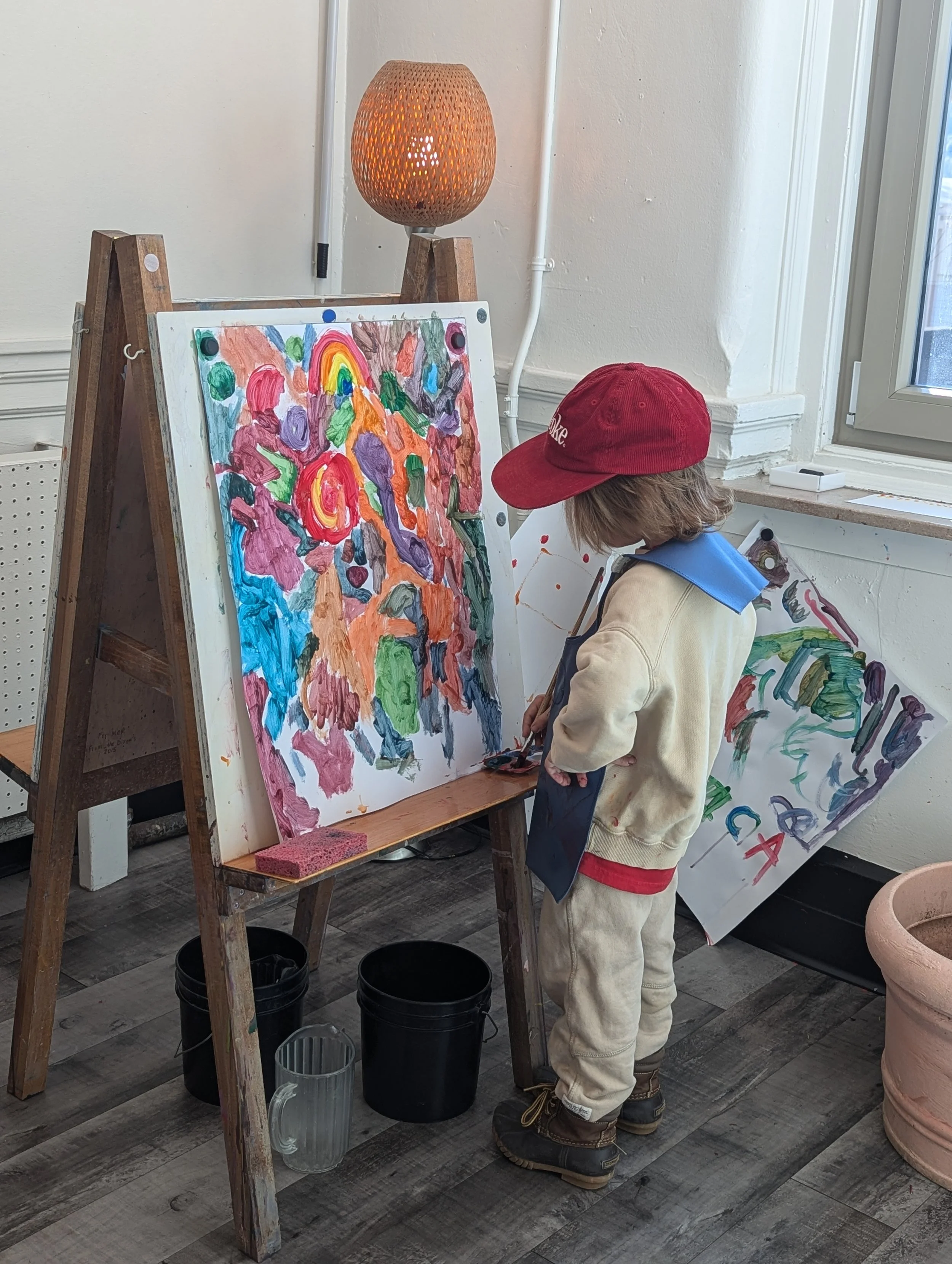 Child standing in front of an easel painting an abstract colorful picture, wearing a red cap, beige sweatshirt, and blue apron, in a bright indoor space with wooden flooring and a window.
