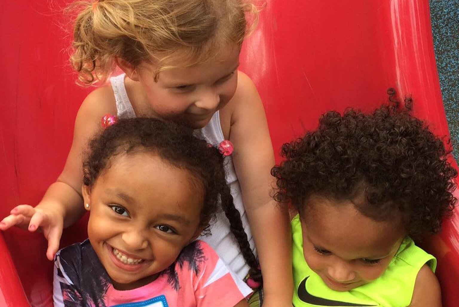Three children riding in a red amusement park ride. One girl with curly blonde hair, another girl with dark curly hair smiling, and a boy with dark curly hair looking down.