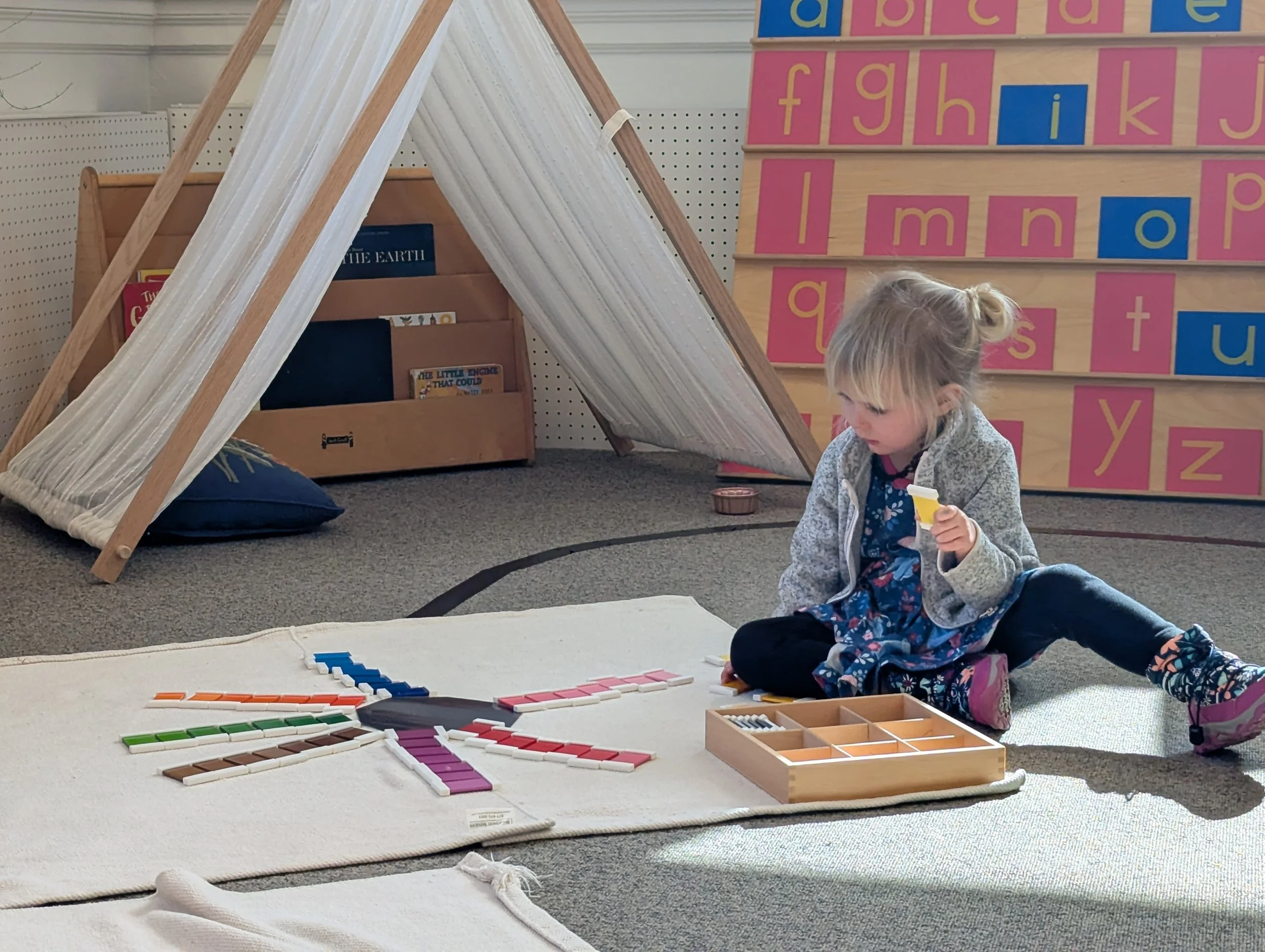 A young girl sitting cross-legged on a carpeted floor, playing with an educational tile set that spells a rainbow on a white mat in a classroom.