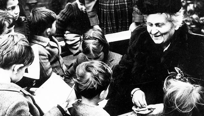 A group of children gathered around a woman with curly hair, smiling and engaging with her, possibly in a classroom or public setting.