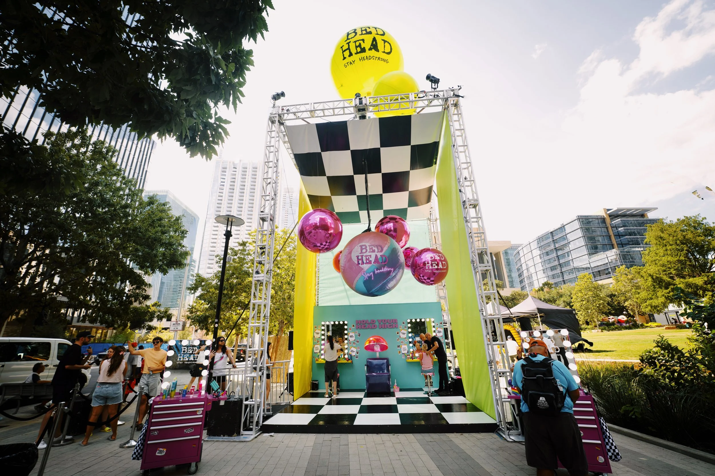 Outdoor event setup with a large yellow inflatable balloon reading "Bed Head. Stay Headstrong." at the top, a checkered black and white canopy, hanging pink and multicolored balloons with "Bed Head" texts, and a mirror station with people getting hair styled. There are trees, modern buildings, and several people around.