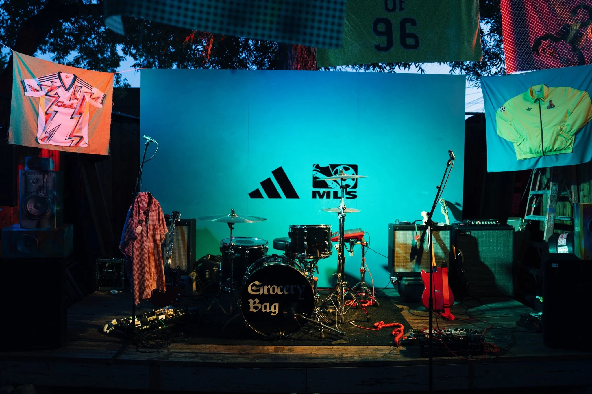 Music stage with drums labeled "Grocery Bag", guitar, amplifiers, microphones, and clothing hanging on either side, including a neon green jacket and a shirt with a lightning bolt design, illuminated with colorful lighting