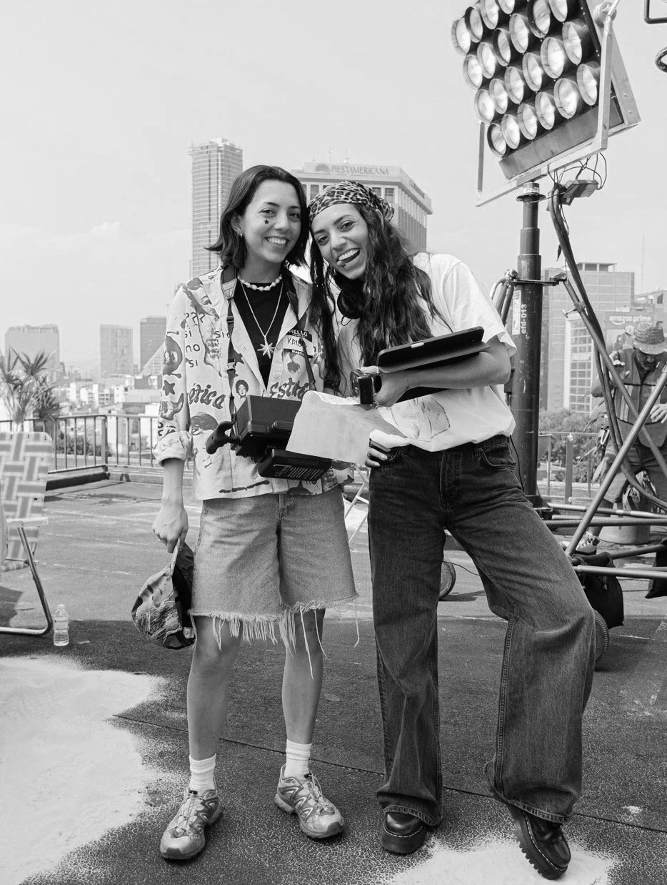 Two women standing together on a rooftop with city buildings in the background. They are smiling and holding documents. One woman wears a patterned shirt, shorts, and sneakers. The other wears a t-shirt, wide-leg jeans, and a bandana. There is large lighting equipment to the right.