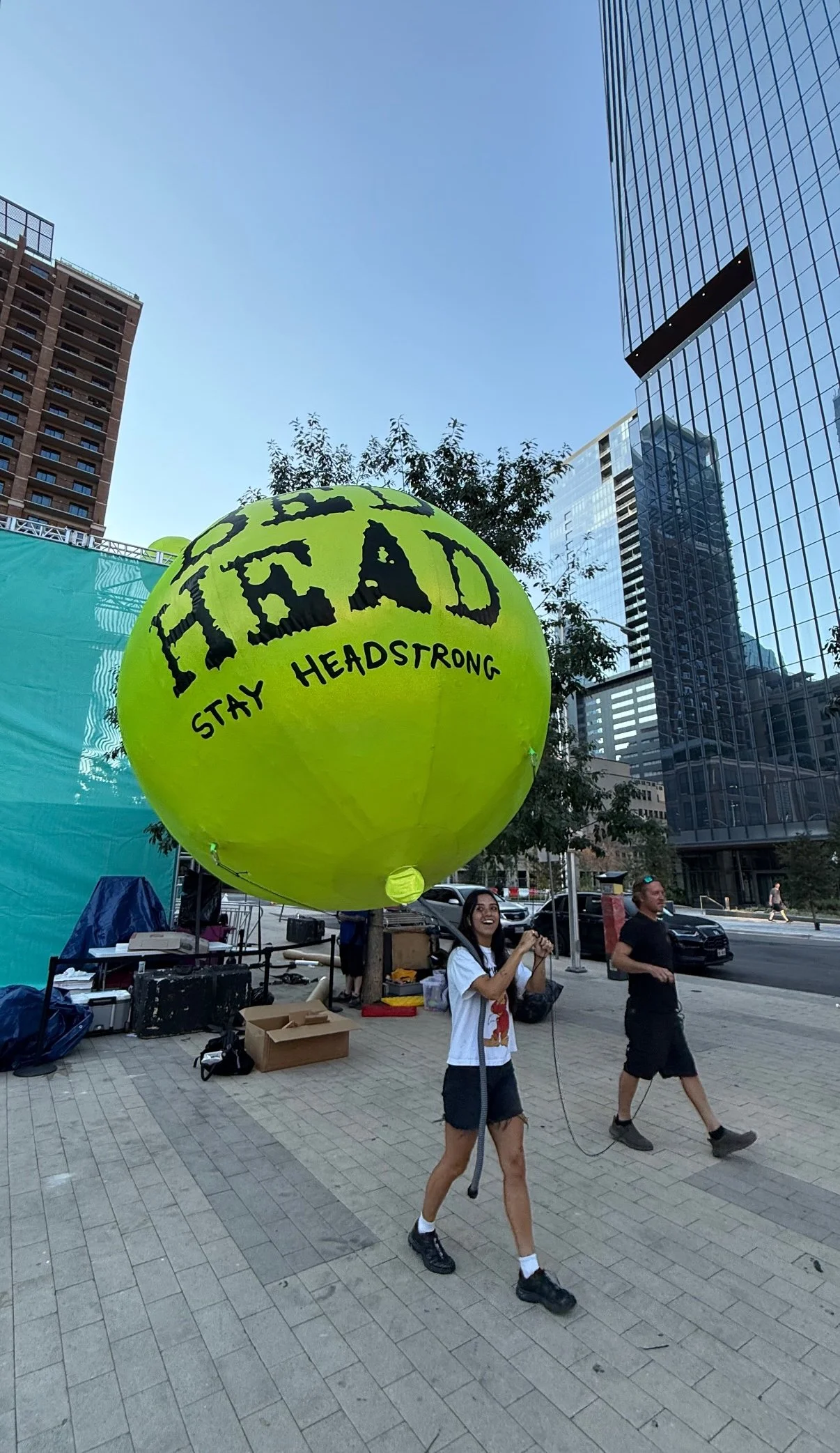 A woman holding a yellow balloon with the words 'HEAD STAY HEADSTRONG' written on it in black letters. She is smiling and walking on a city sidewalk with tall buildings in the background.