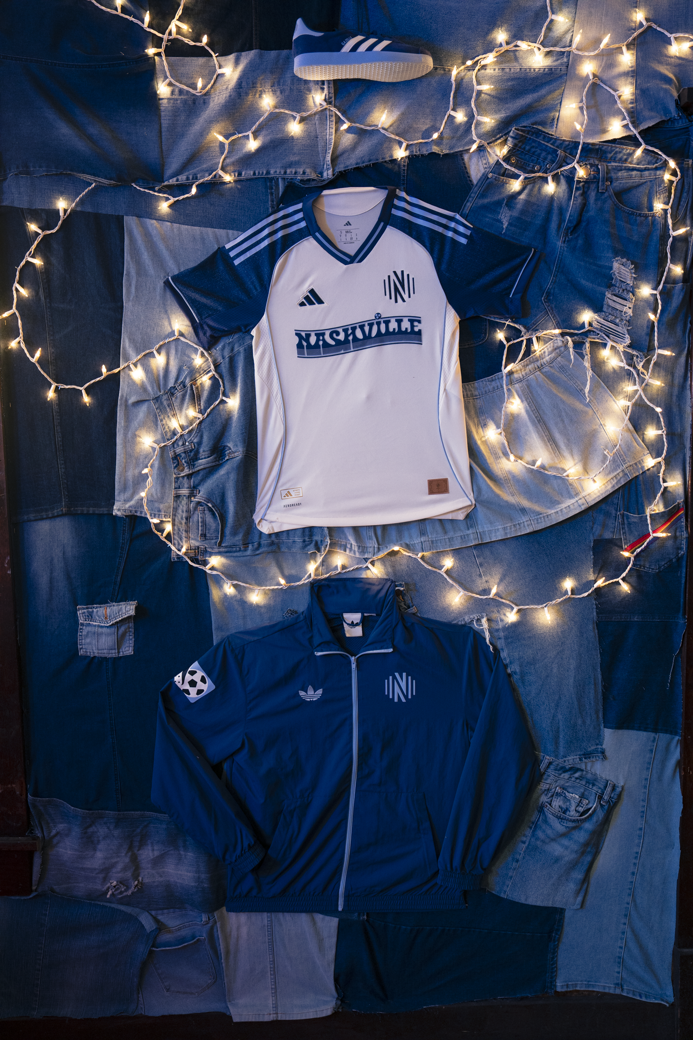 Display of two Nashville soccer jerseys, one white and one navy blue, placed on layered denim patches with string lights around them.