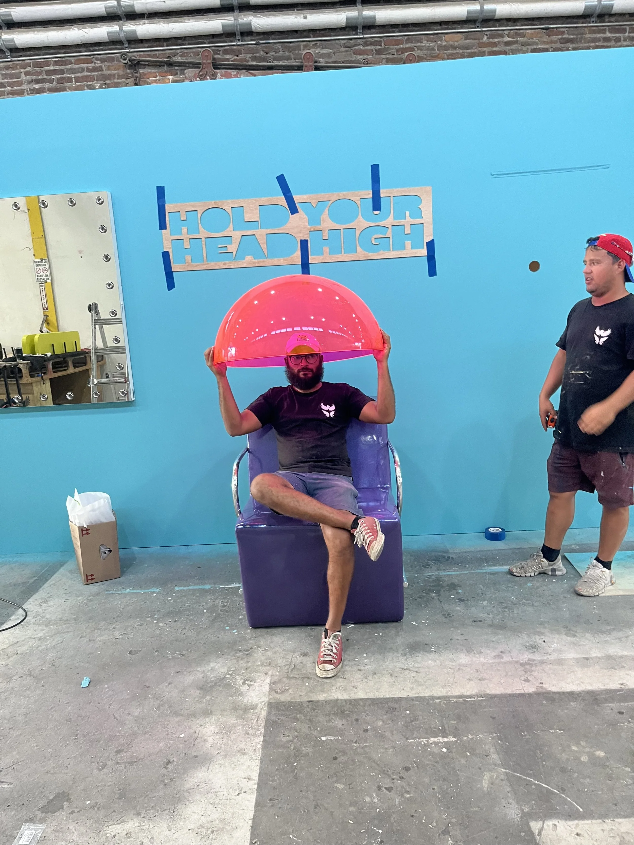 Man sitting on a purple chair holding a large red exercise ball over his head inside a gym with a blue wall behind him that says "Hold your head high". Another man stands to the right, wearing a black t-shirt and shorts, looking at him.