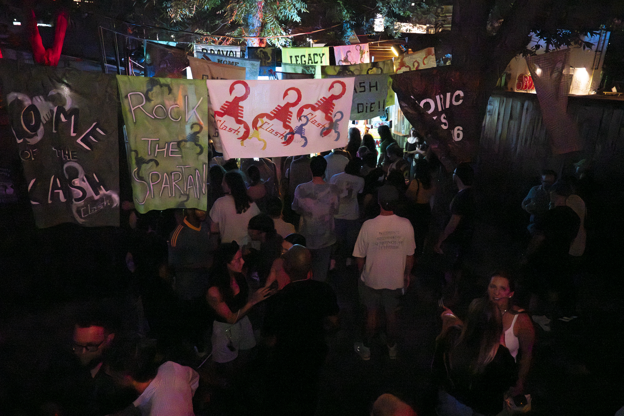 People gathered outdoors at night with colorful banners and flags hanging above them, celebrating or attending an event.
