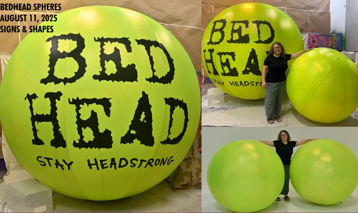 Image of three large green spherical objects resembling balls, with one prominently displaying the text "BED HEAD STAY HEAD STRONG." A woman is seen interacting with the spheres; in one image, she is smiling and touching one sphere with her arm resting on another, and in another, she is standing between two spheres with arms stretched out to the sides. The background has a light-colored wall and some abstract art or signs.