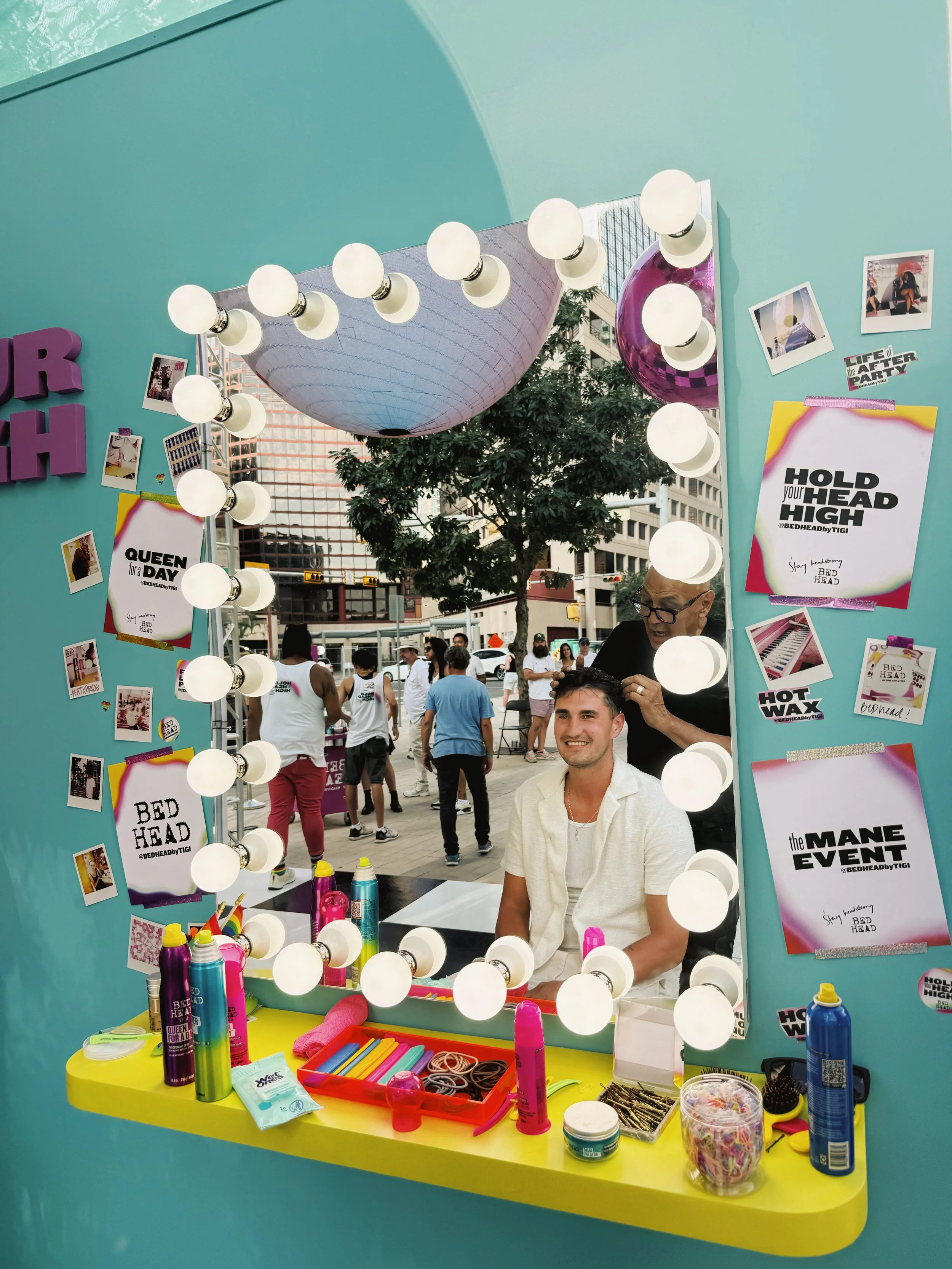 A young man getting a haircut in front of a large mirror with round lights around its frame. The scene is outdoors with several people in the background. The mirror has colorful signs and Polaroid photos taped around it, and a yellow table with hair products and accessories in front of it.