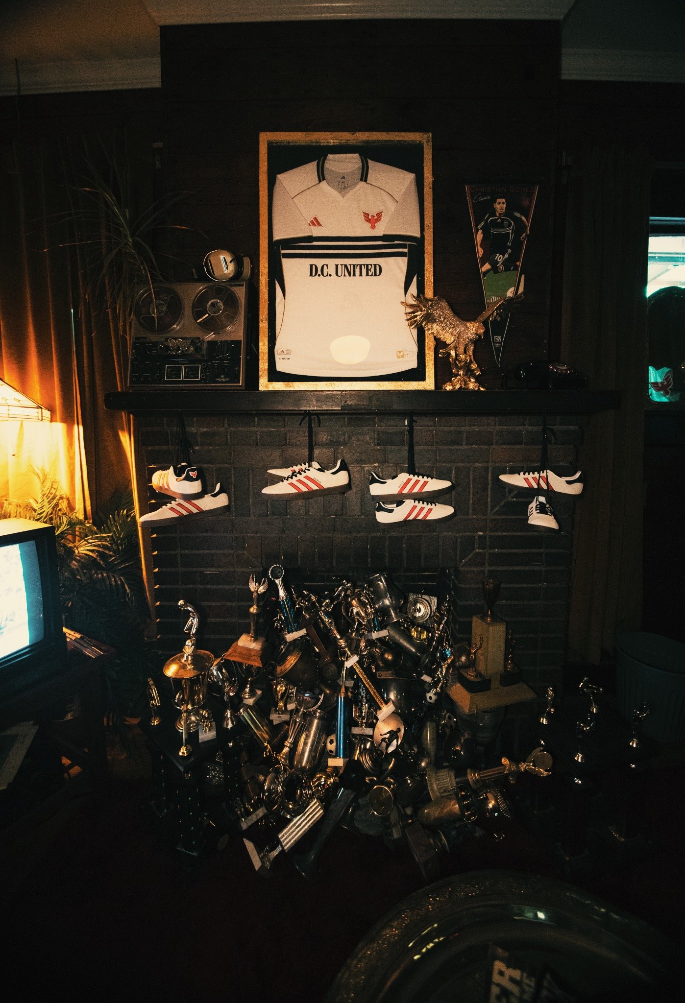 A display with a framed D.C. United soccer jersey on a dark brick wall, surrounded by sneakers, trophies, and various metallic items, with a vintage stereo and a small television on either side.