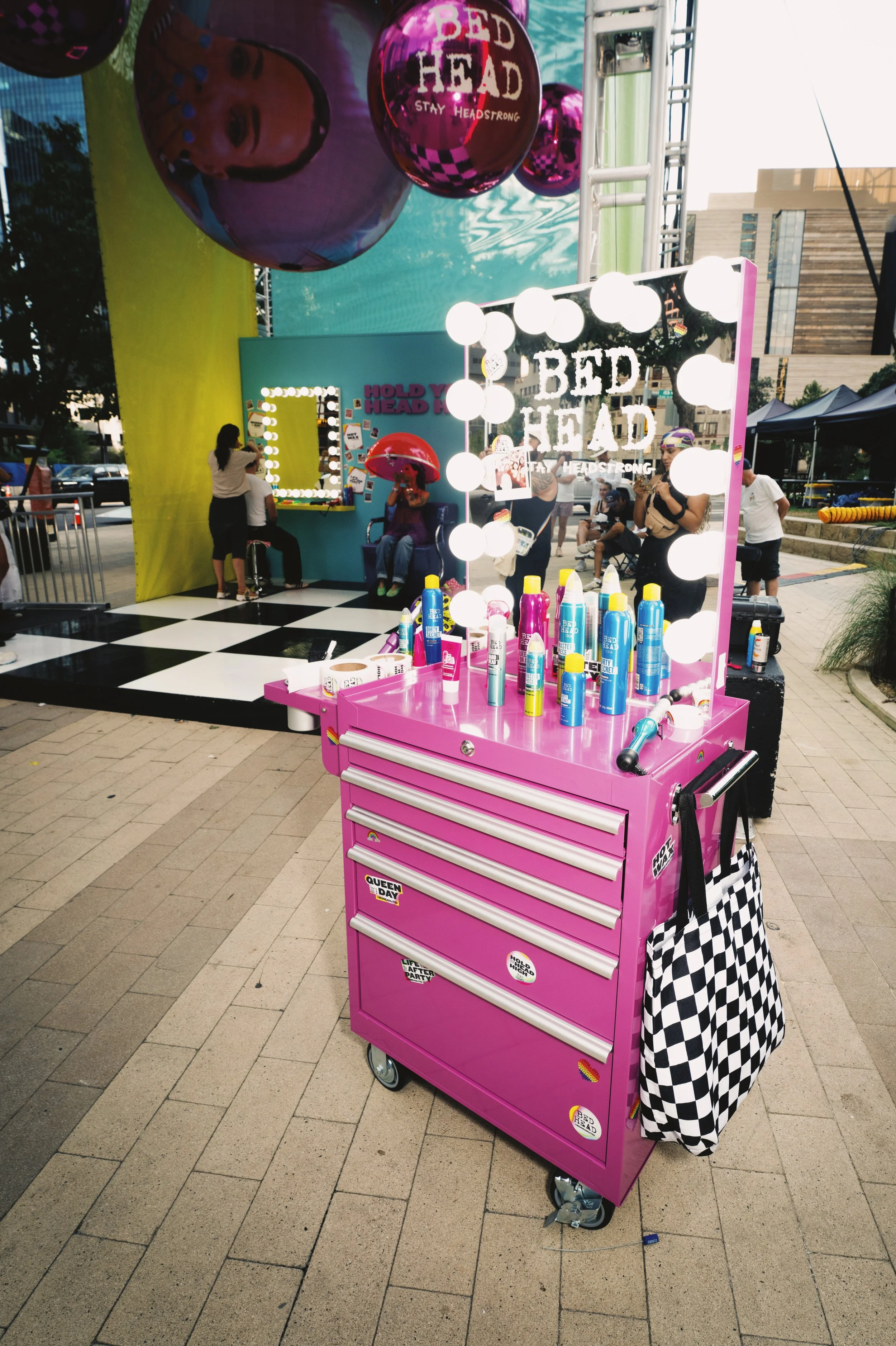Pink rolling tool cart with hair spray bottles and a checkered bag, illuminated mirror with round light bulbs, and a sign reading 'BED HEAD' in the background, at an outdoor event with people and festive decorations.