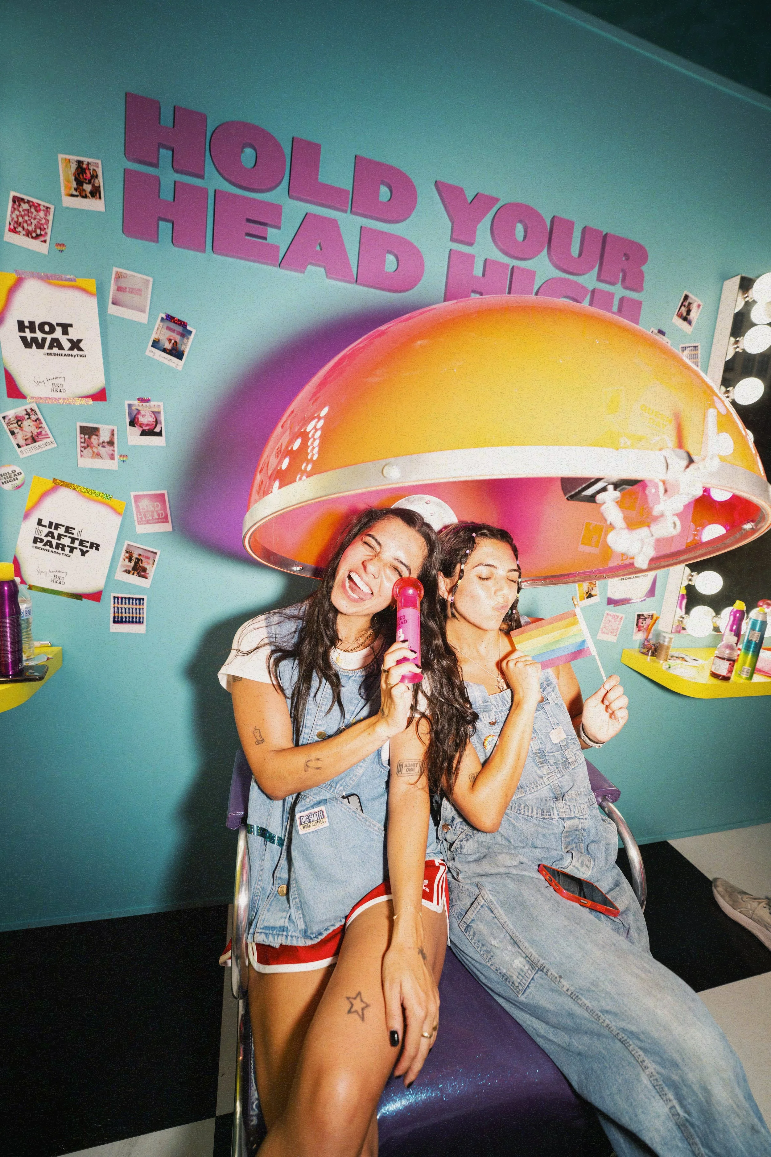 Two young women sitting under a large pink and yellow lamp with a backward rainbow flag, in a colorful, lively room with Polaroid photos on the wall and the phrase "HOLD YOUR HEAD HIGH" in large purple letters on the wall.