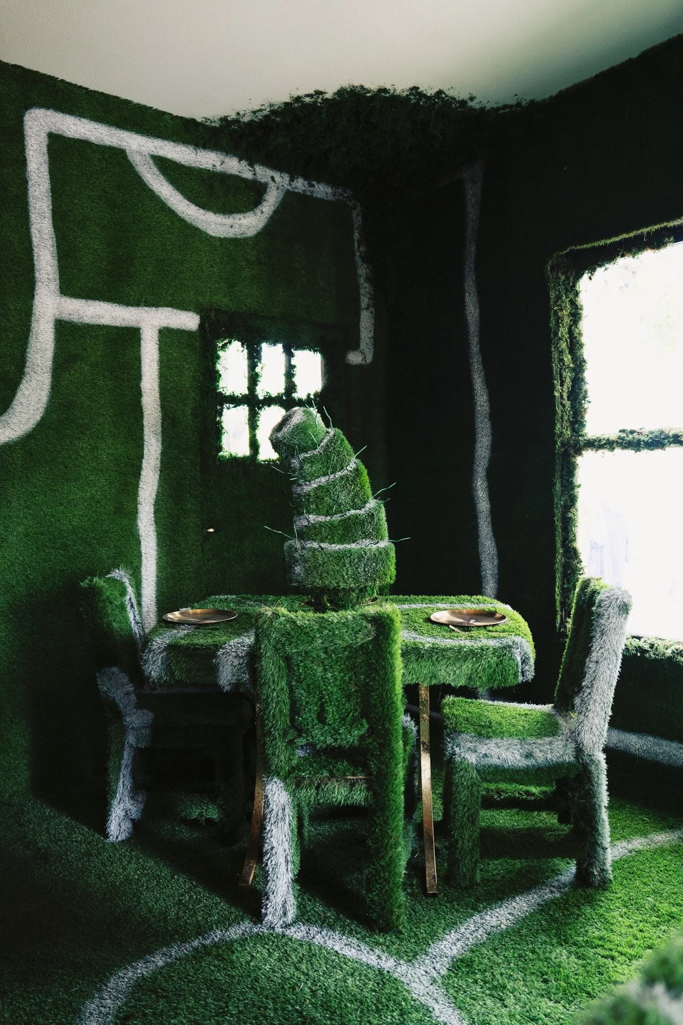 Room decorated with green and white grass-covered furniture including chairs, a table, and a centerpiece, with walls and floor also covered in grass creating an indoor garden look.