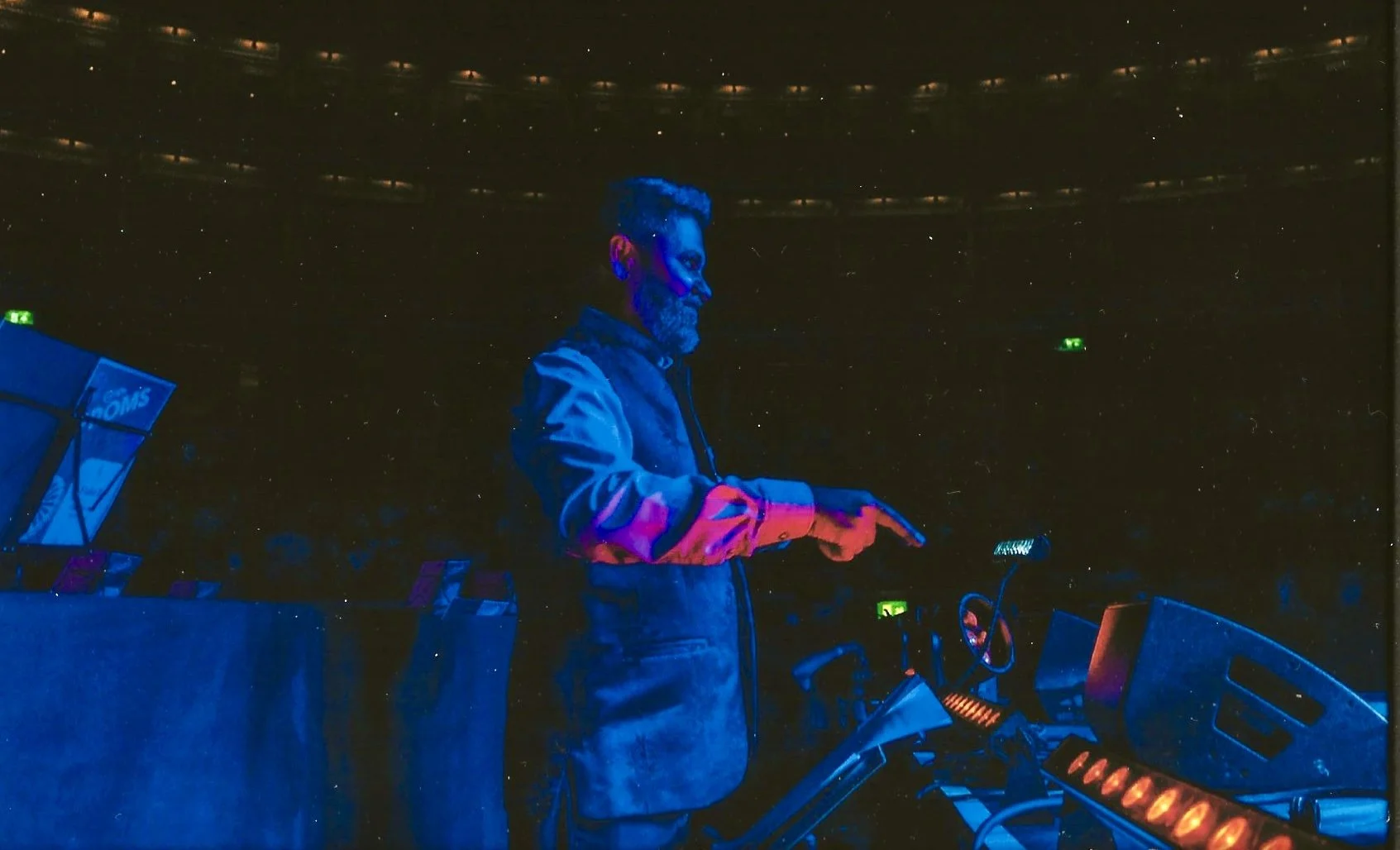 A DJ with blue and purple face paint, wearing a jacket, performing at a dark music event, with DJ equipment and a motorcycle in front of him.