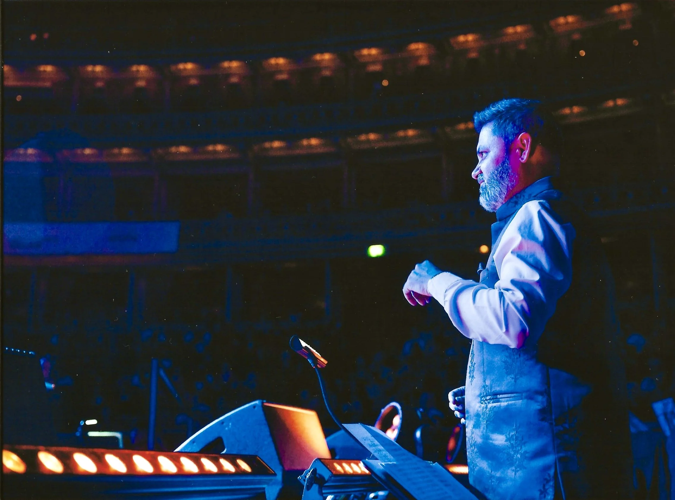 A man with a beard standing on a stage in a theater, illuminated by stage lights, with a music stand and sheet music in front of him.