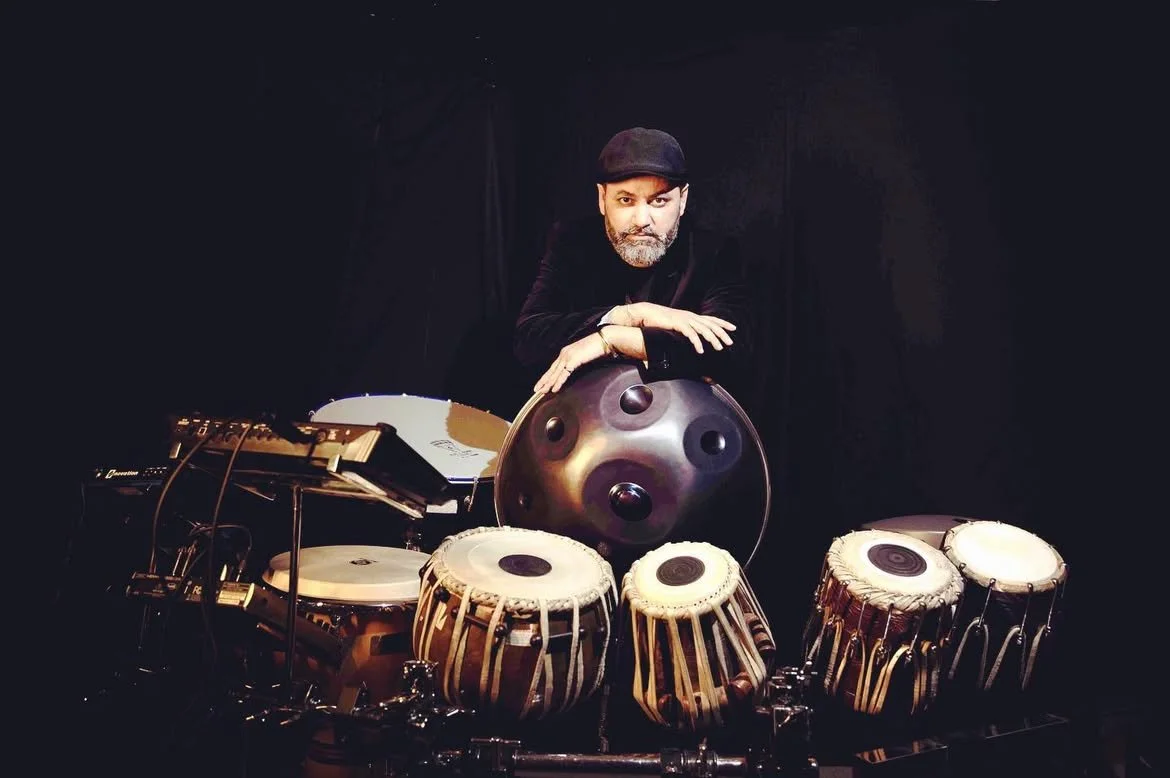A man with a beard wearing a black cap and dark clothing is standing behind a table of musical instruments, including tablas, a hang pan, and electronic equipment, in front of a black background.