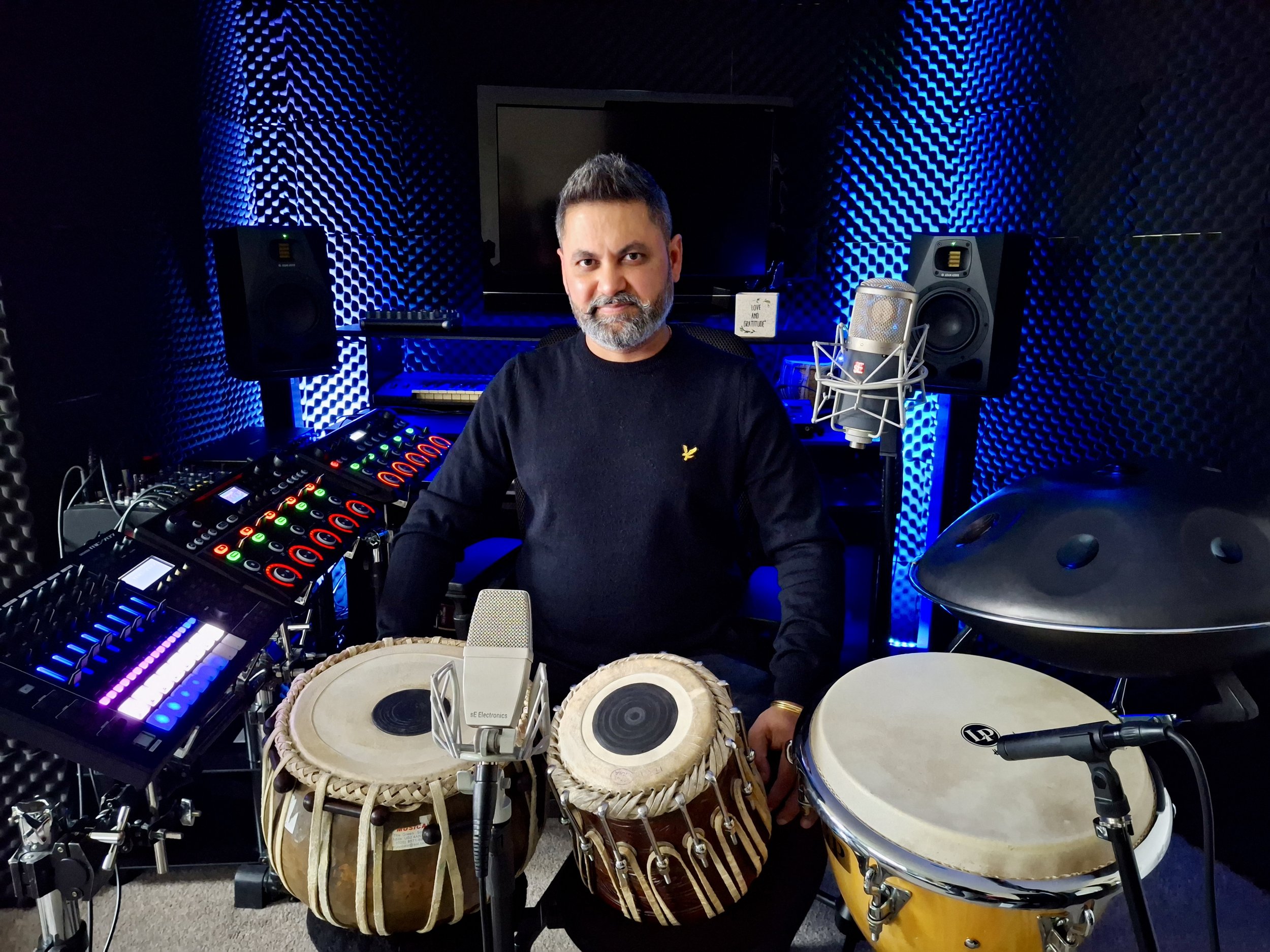 A man with a beard and short hair sitting in a music studio surrounded by musical instruments and equipment, including two drums, a keyboard, speakers, and a microphone, with acoustic foam panels on the walls and a black monitor screen in the backgro