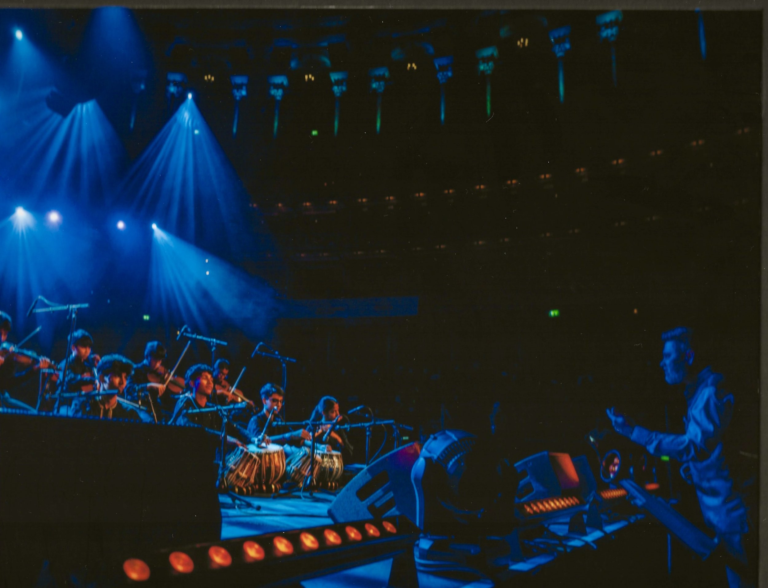 A group of musicians playing traditional instruments on stage, with a conductor or sound engineer at a console to the right. The scene is lit with blue stage lighting, in a dark auditorium with visible ceiling lights.