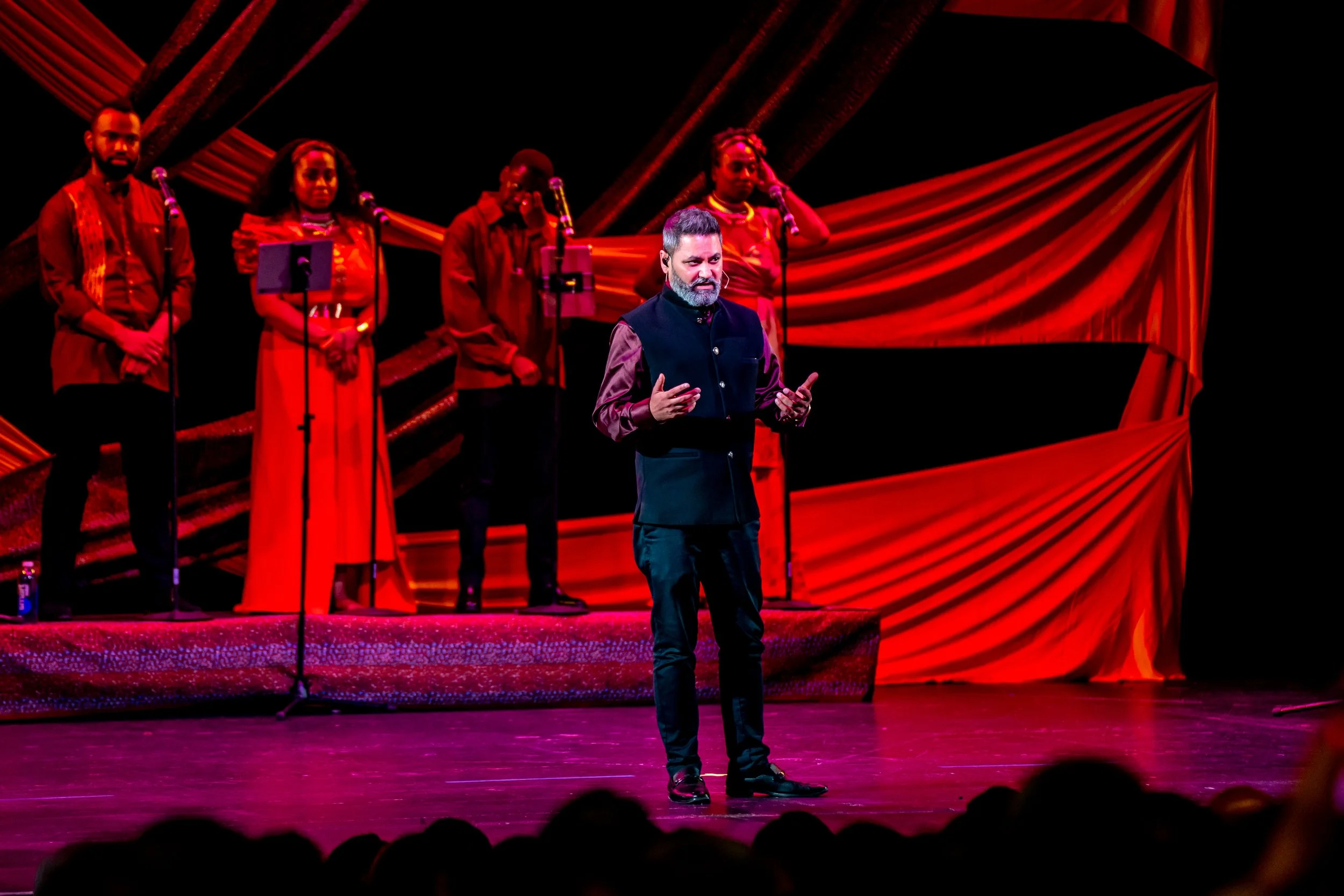 Man performing on stage with a group of singers in the background, decorated with red curtains and stage lighting.