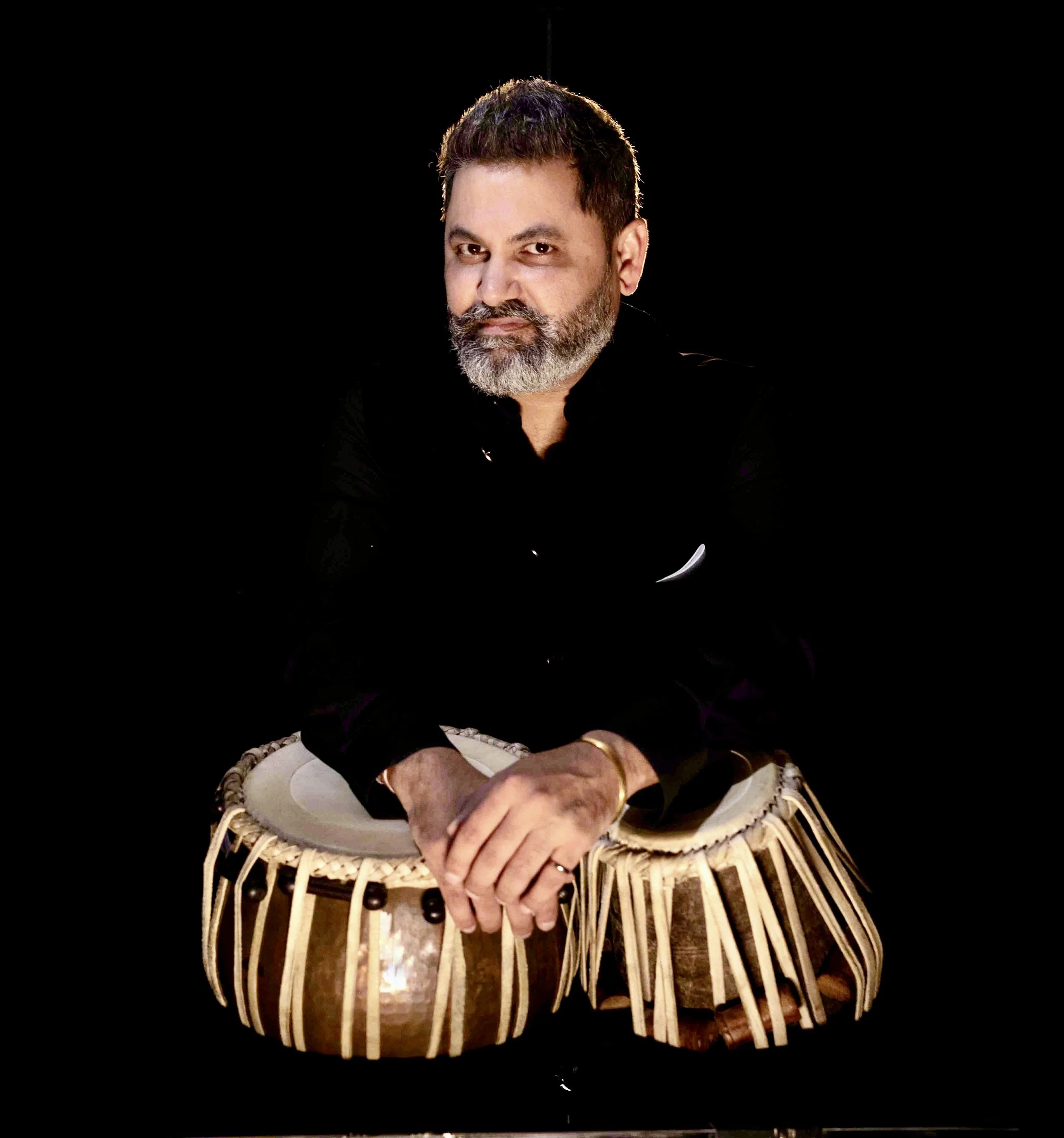 A man with a beard dressed in black, sitting with hands resting on a tabla against a black background.