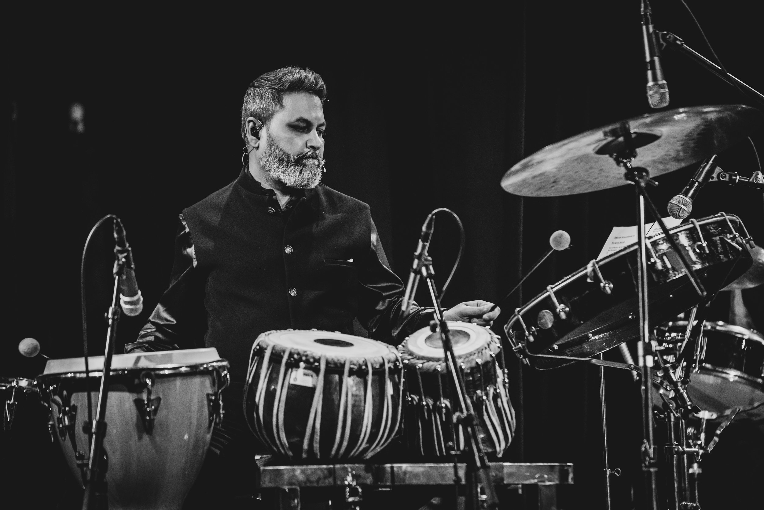 Black and white photo of a male musician with a beard playing percussion instruments on stage, surrounded by microphones and drums.