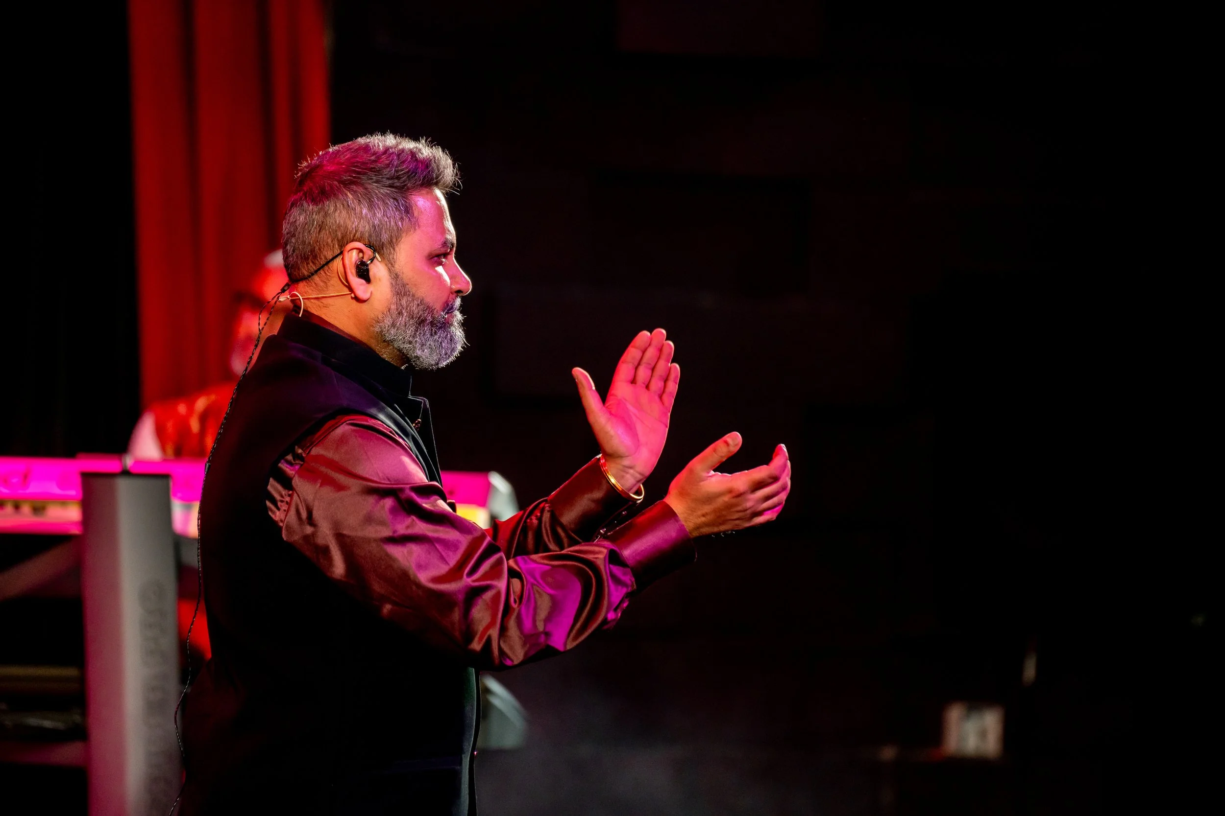 A man with gray hair and beard, wearing a dark vest and a brown long-sleeve shirt, is standing with his hands raised, speaking or singing in a dark room with stage lighting.
