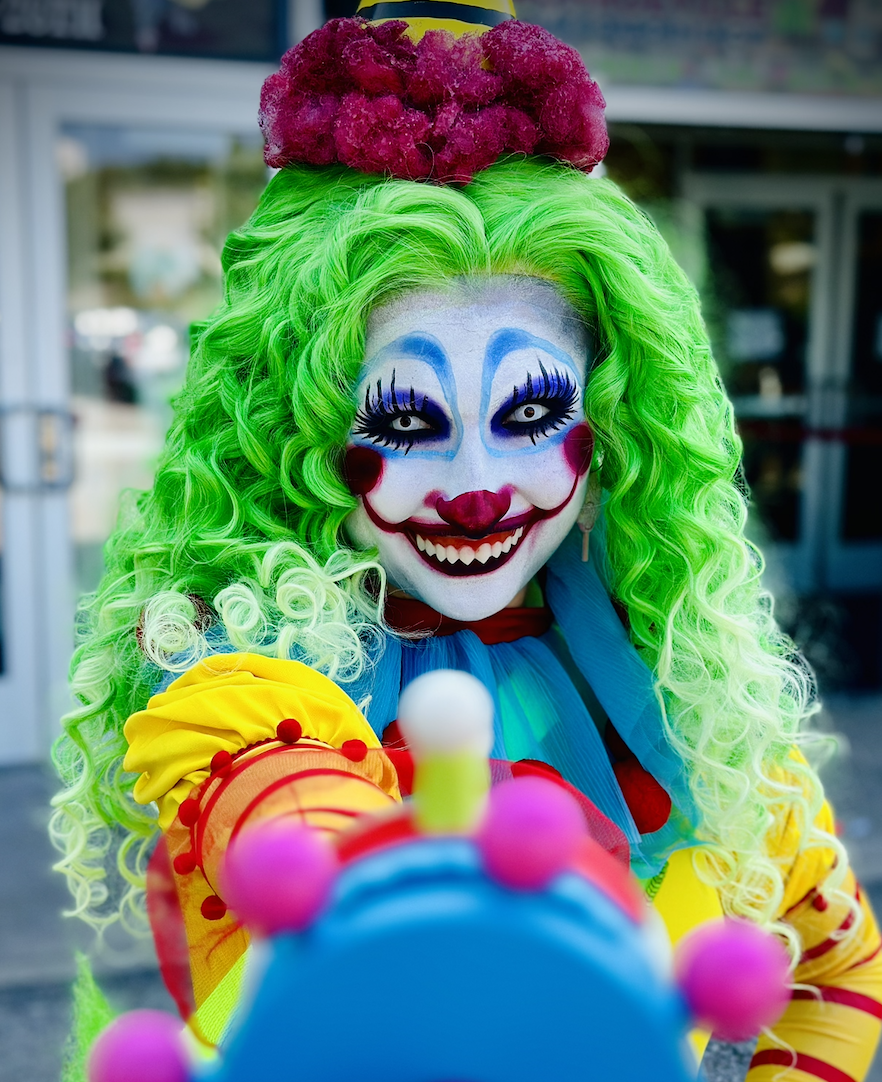 A person dressed as a clown with a colorful costume, bright green curly hair, and clown makeup, smiling and pointing a finger forward.