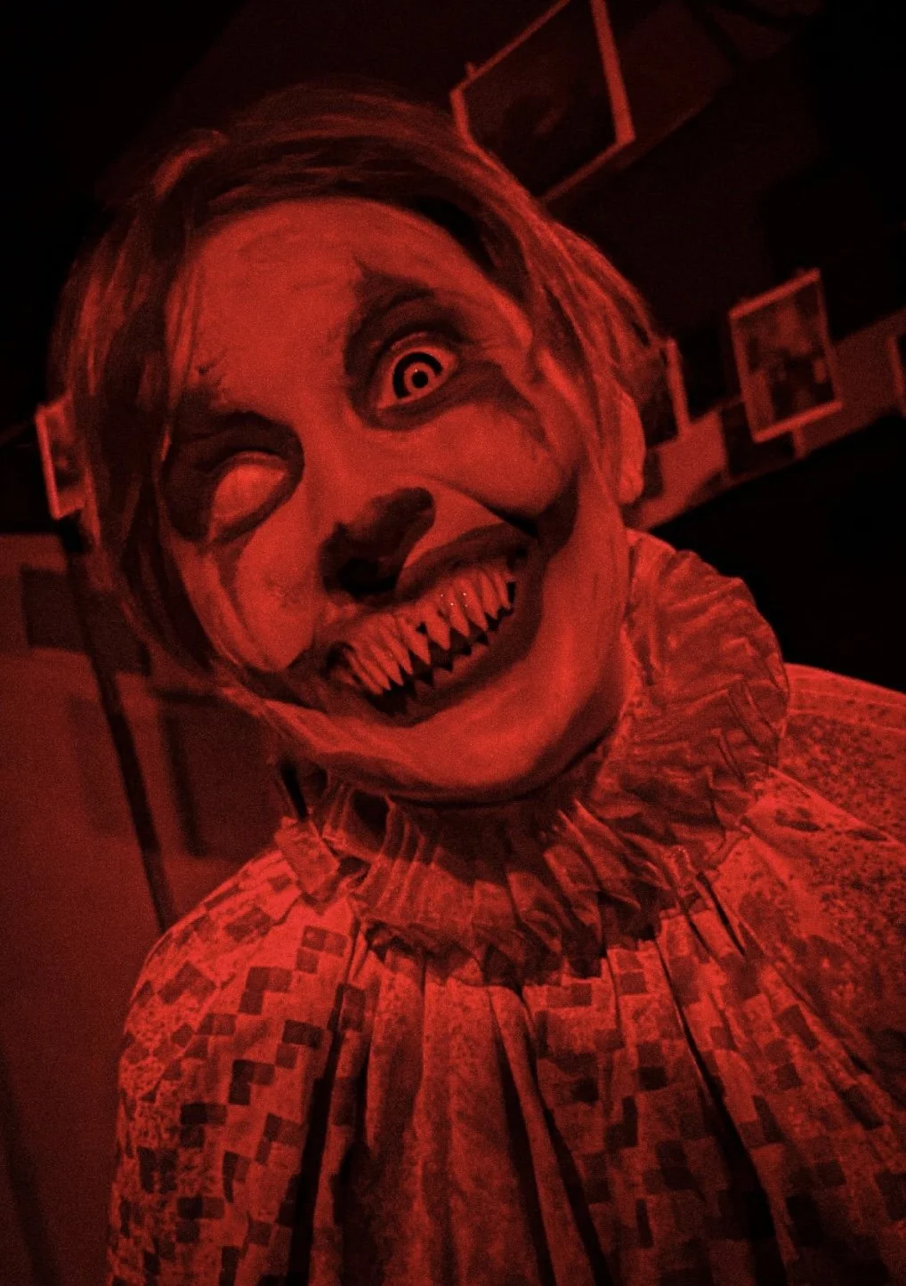 A person wearing a creepy clown mask with sharp teeth and dark eye makeup, illuminated by red lighting.
