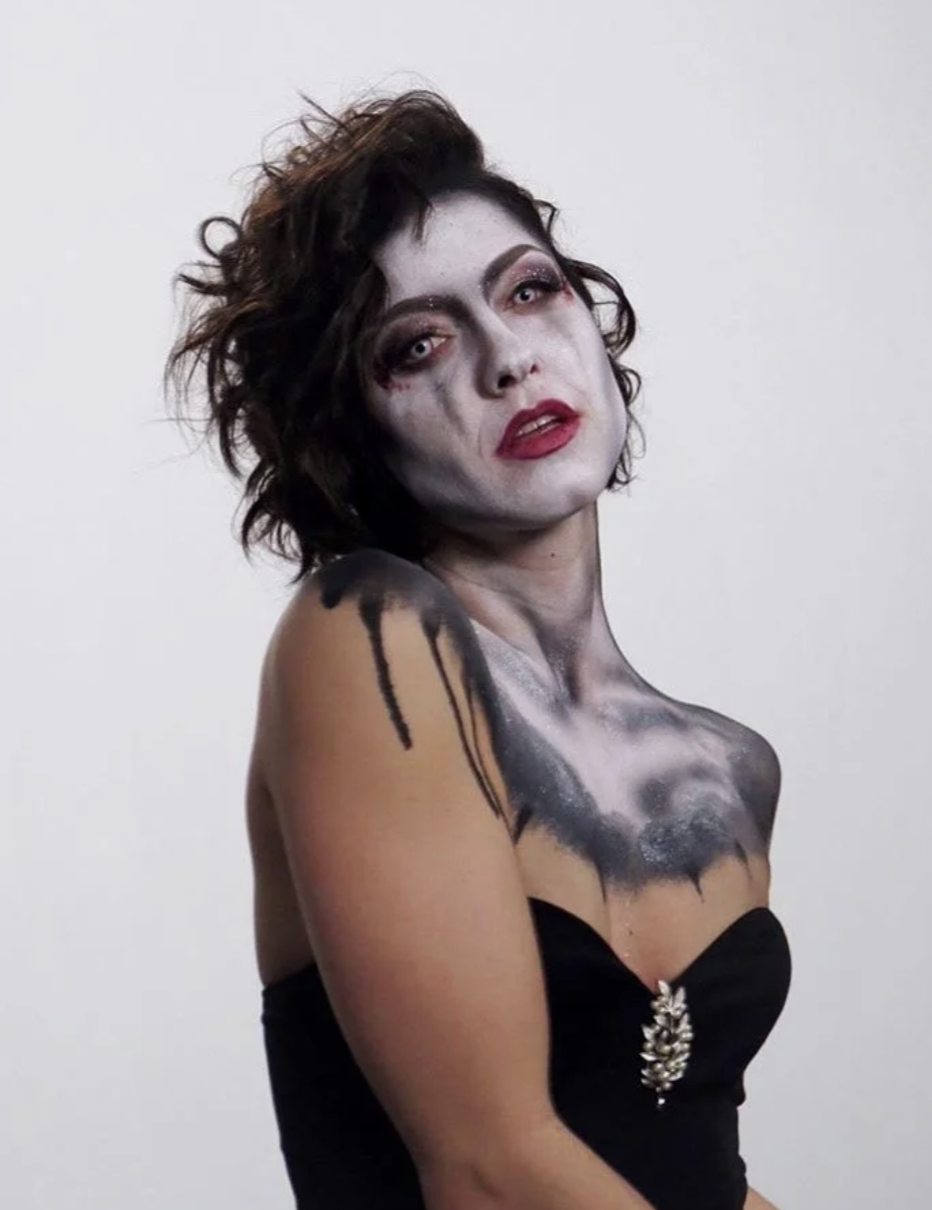 A woman with short, messy dark hair and pale makeup, wearing bold makeup including red lipstick and pink eyeshadow, sports a dramatic look. She has a black strapless top with a decorative brooch and a black and gray painted design on her upper chest 