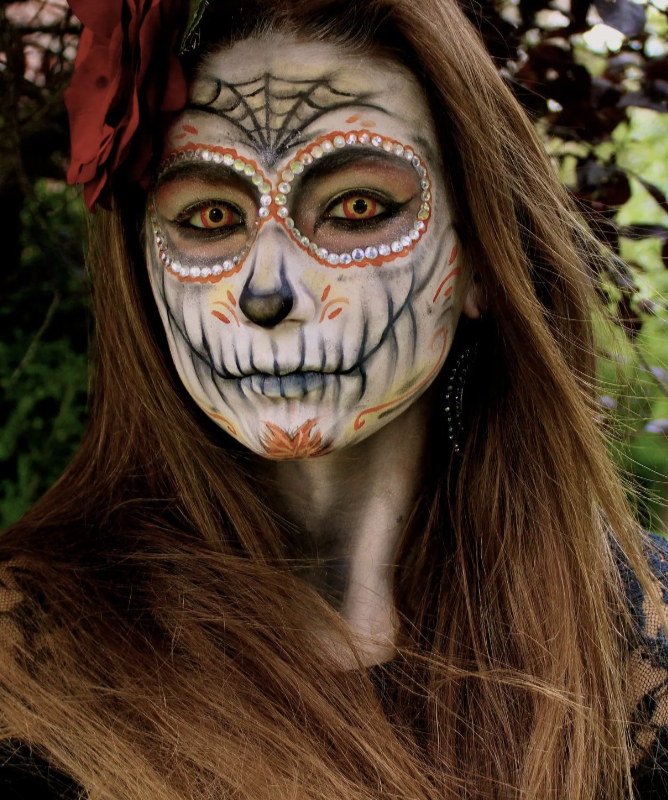 Woman with face painted as a sugar skull, featuring detailed eye designs, a black nose, and orange and white decorative patterns, in a setting with green foliage.
