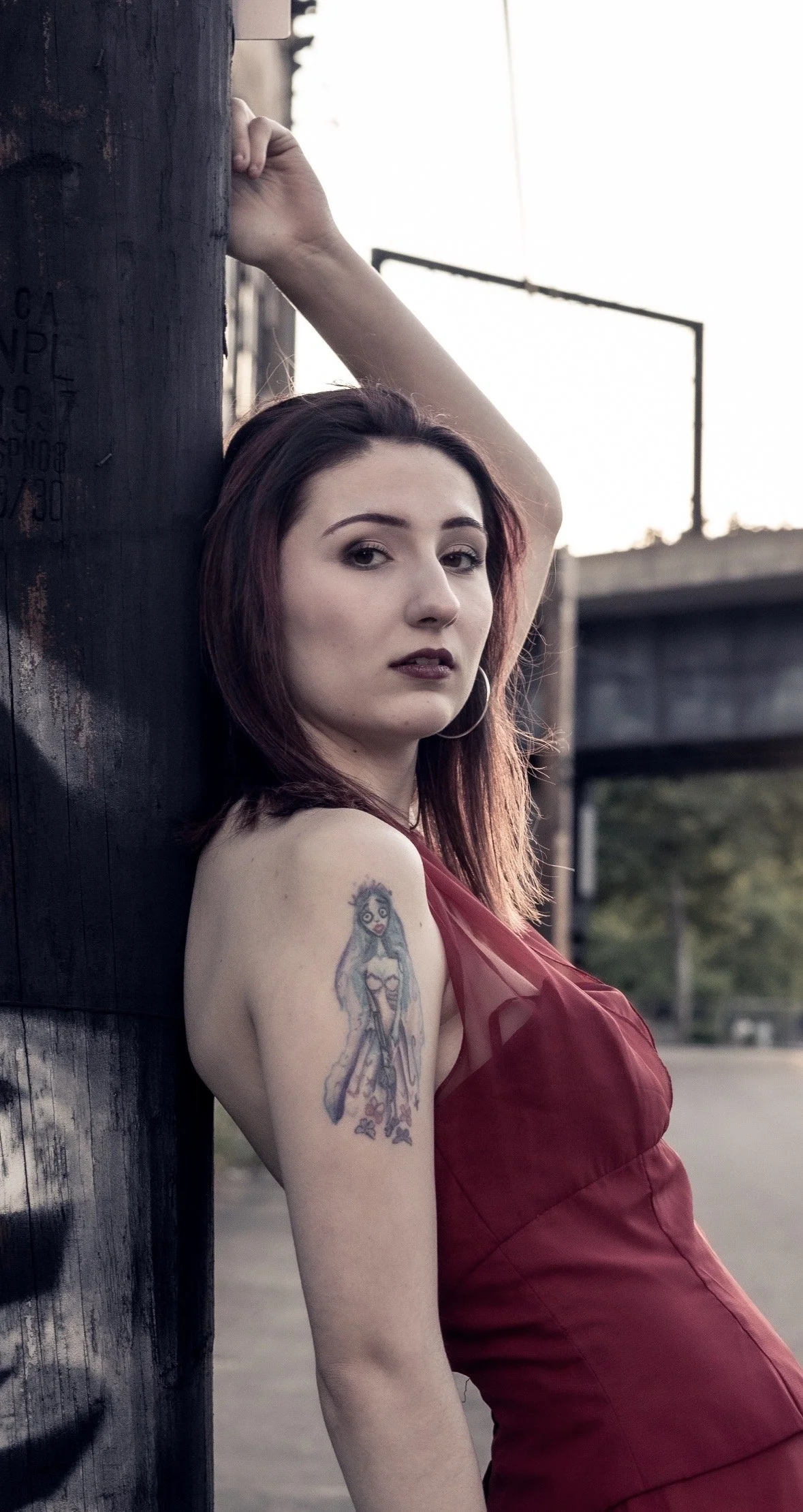 A woman with shoulder-length red hair, wearing a red dress, leaning against a wooden pole outdoors, with a tattoo of a mermaid on her upper arm, and an intense expression on her face.