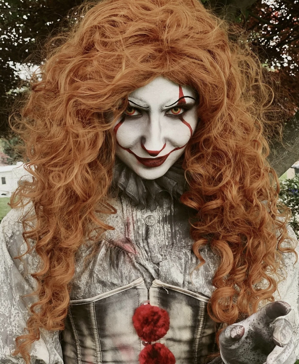Person dressed as a sinister clown with white face paint, red markings, and curly red hair, outdoors.