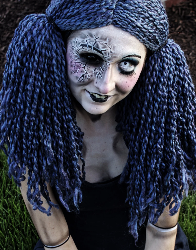Person with long, thick, blue braided hair and gothic makeup, including cracked face makeup around the eye and dark lipstick, sitting outdoors on grass.