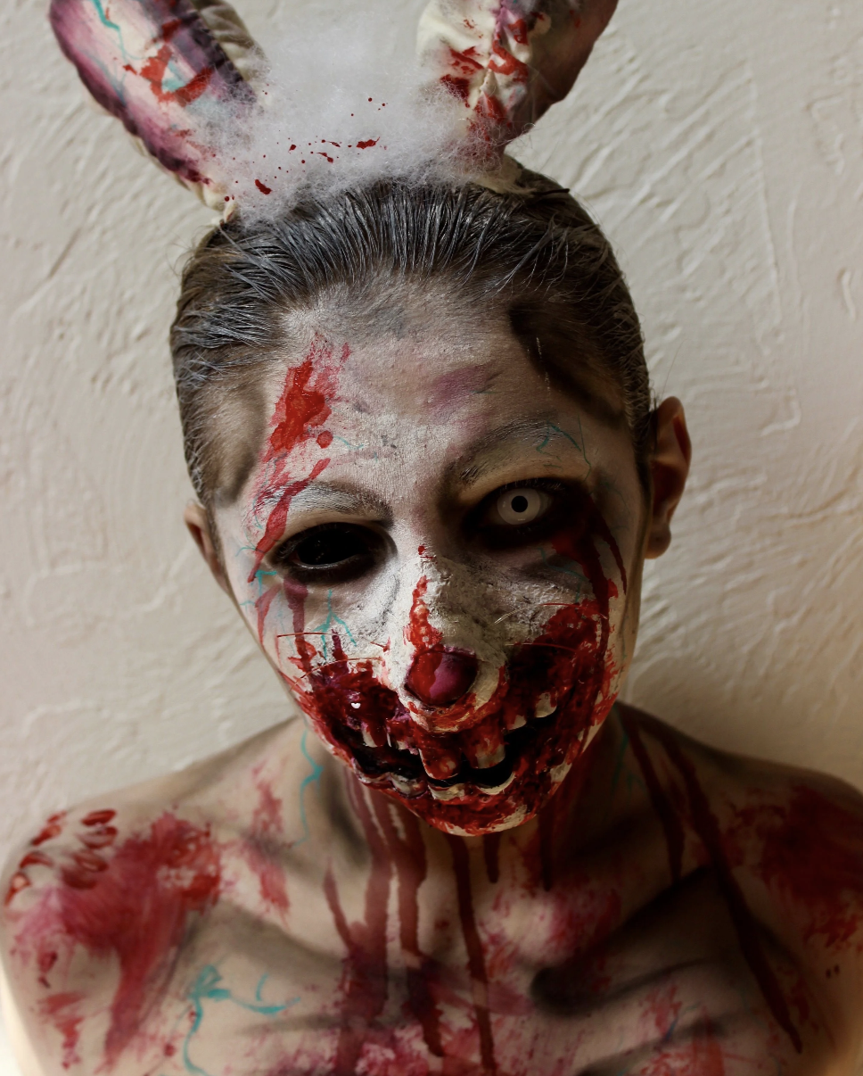 A person dressed as a creepy clown with distorted white face makeup, blood-like red paint, black eyes, and a sinister expression. They have a large, blood-spattered bunny ears headband and are shirtless, with face and body painted and smeared with re