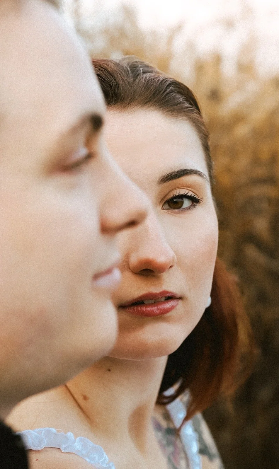 Close-up of a woman with brown hair and makeup, partially obscured face of another woman in foreground, out of focus.