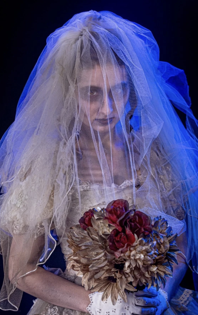 A woman dressed as a bride with a veil over her face, holding a bouquet of dried and artificial flowers, with a dark background and dramatic lighting.