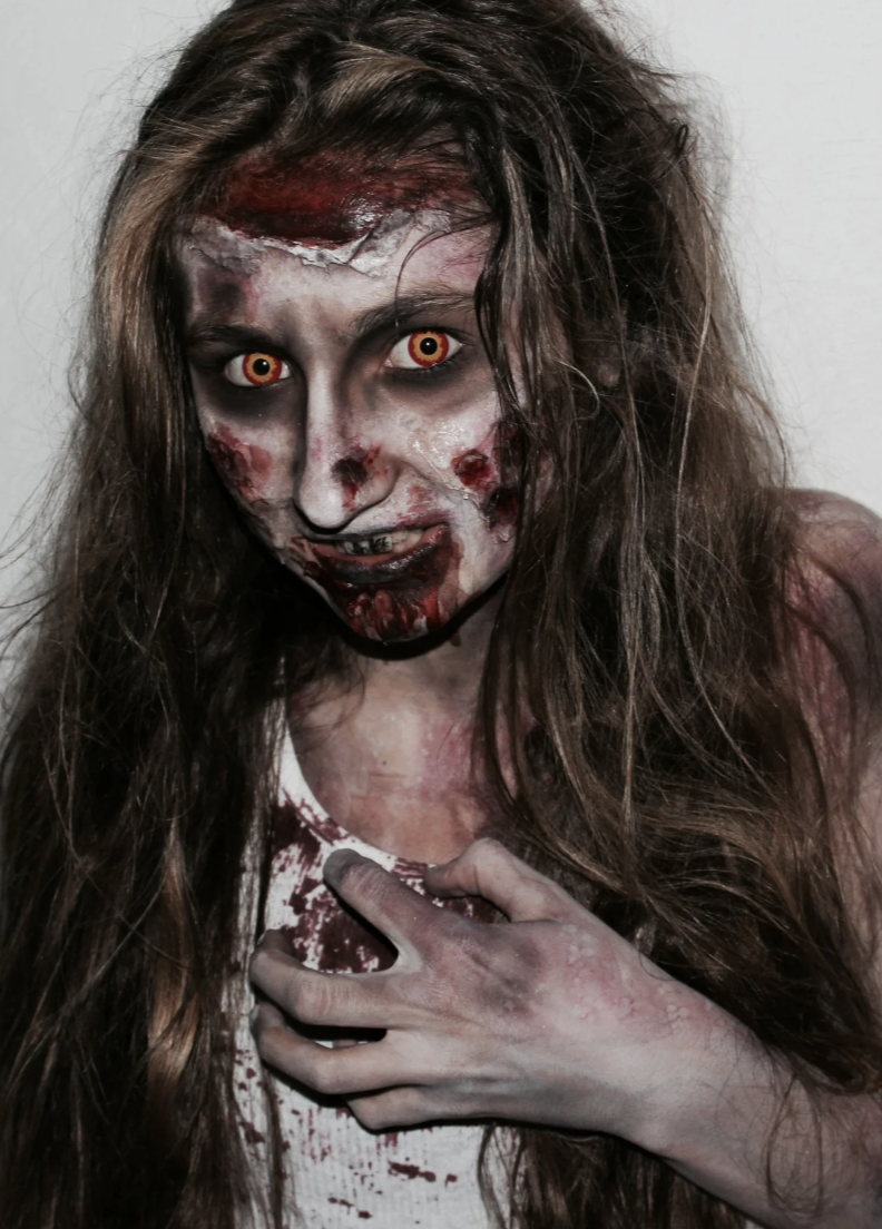 A person with zombie makeup, pale skin, red and black facial scars, glowing yellow-red eyes, messy brown hair, and wearing a bloodstained white tank top.
