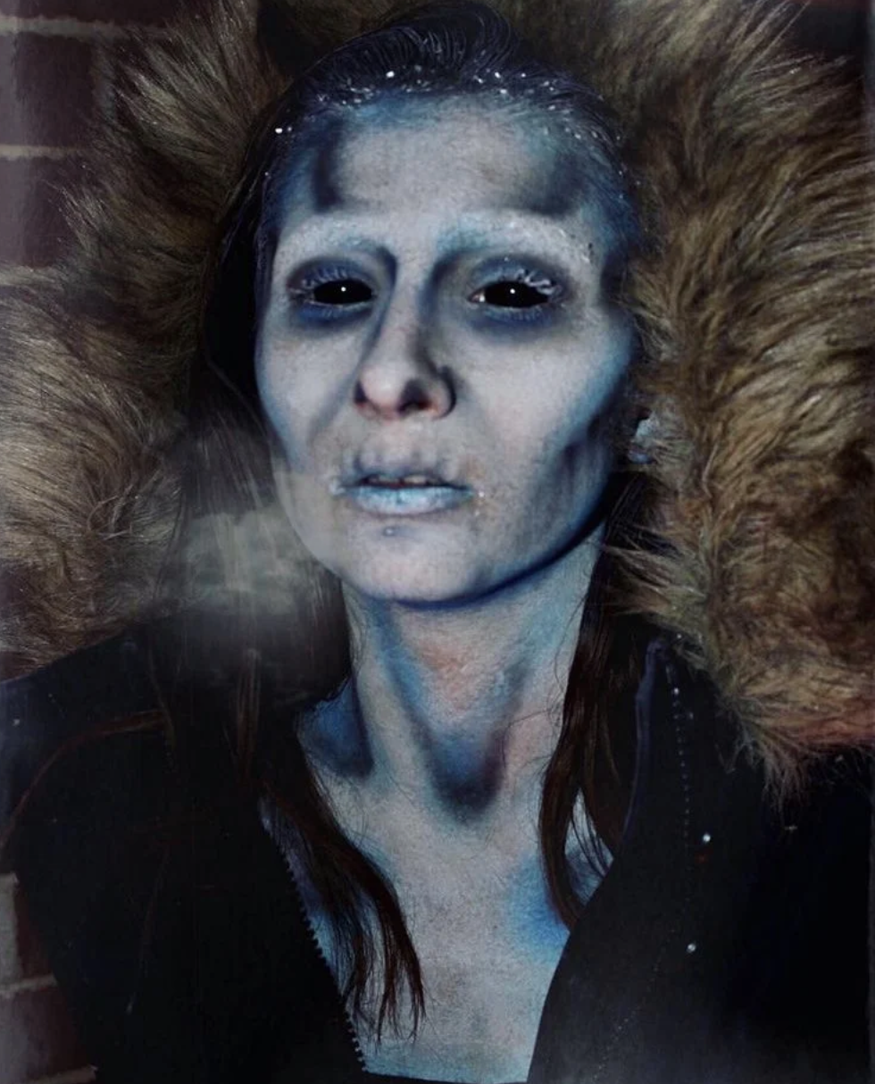 Doll or mannequin with icy blue makeup, black eyes, and long brown hair. It is dressed in a black coat with a fur-lined hood and is behind glass, giving an eerie appearance.