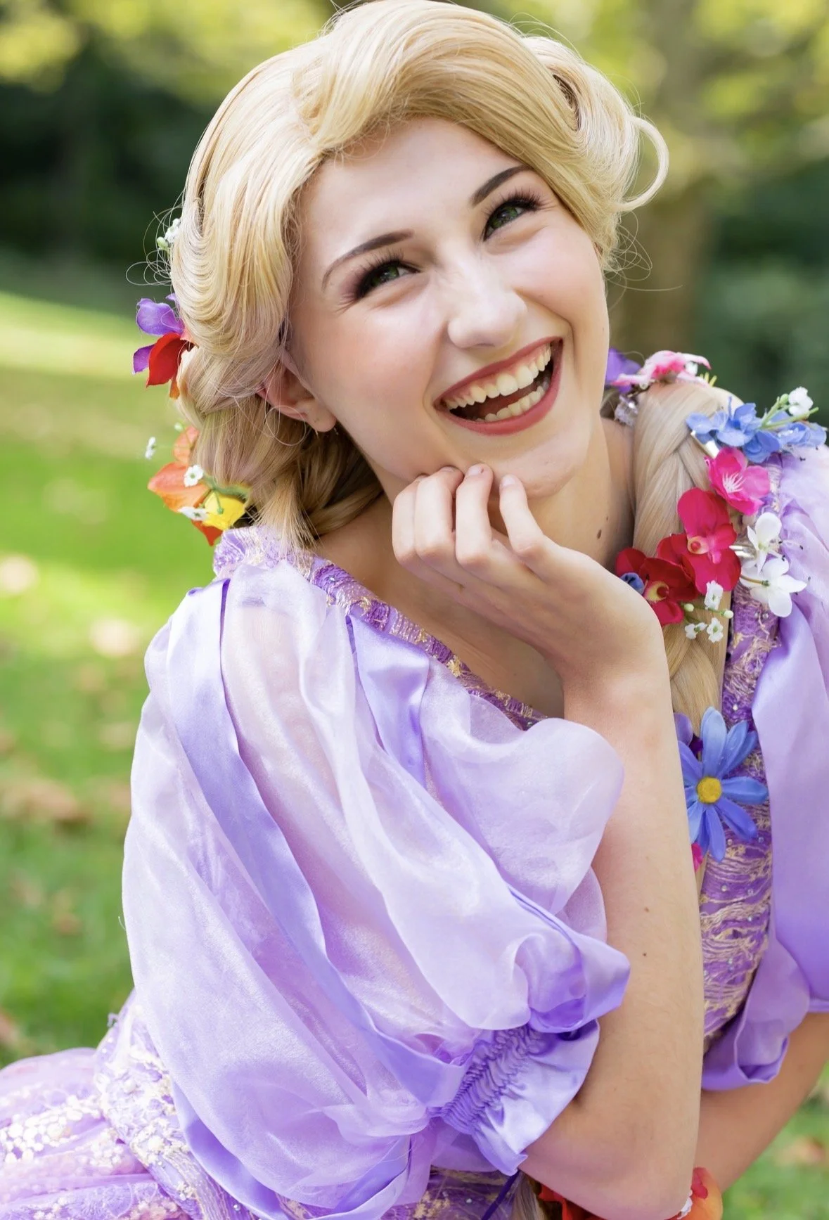 A woman with blonde hair and a joyful expression, wearing a purple dress with puffed sleeves and decorated with colorful flowers in her hair, outdoors with green foliage in the background.