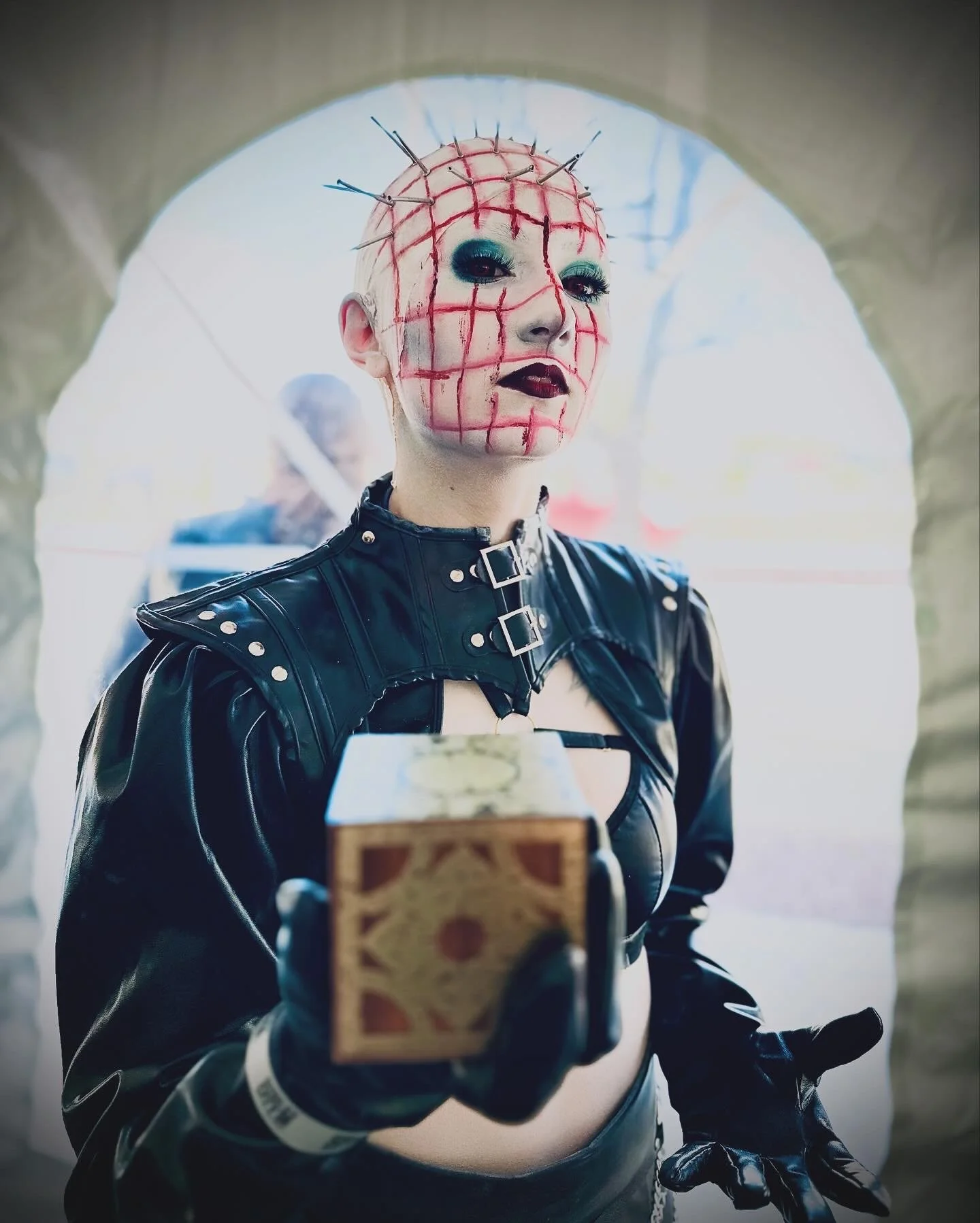 Person dressed in a black leather outfit with face painted in red grid pattern and spikes on scalp, holding a tarot card, standing in front of stone archway.